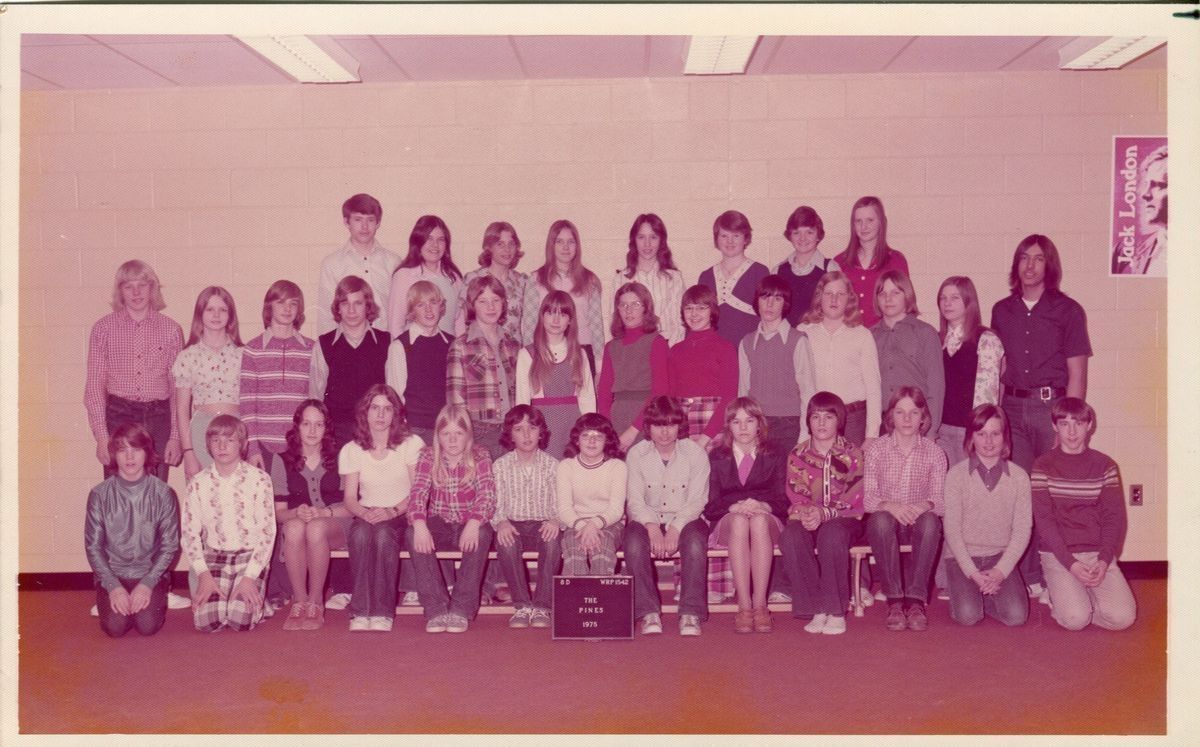 The Pines class photo 1975.: This is a coloured photograph of a class from The Pines of thirty-five students. They are standing in three rows in front of a white brick wall while wearing a variety of 70's fashion. To the right of the group there's a poster of Jack London, and in front of them is a black plaque with white letters that reads '8D WRP1542 The Pines 1975'.