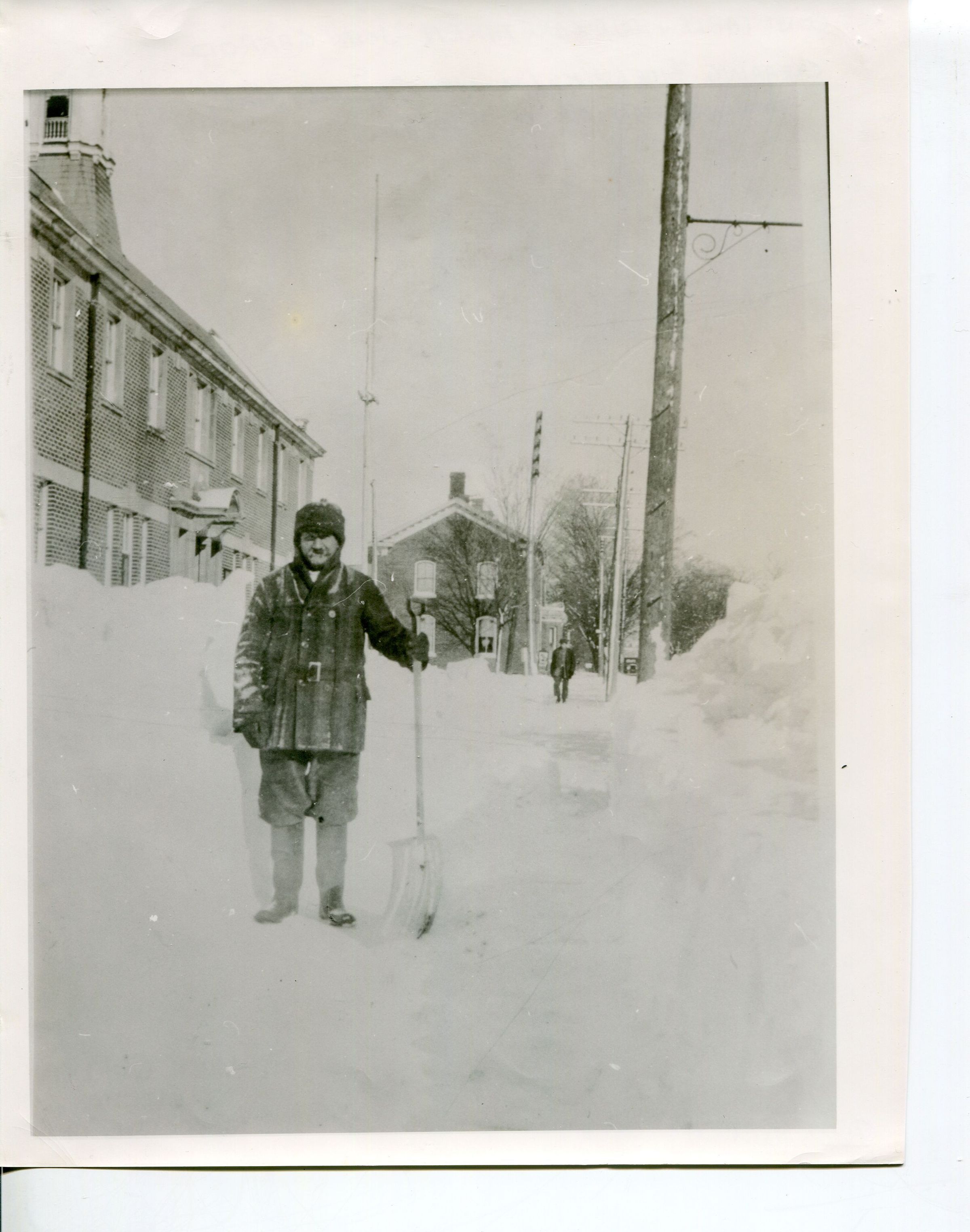 In this black and white photograph from circa 1925, Constable John Garrod is depicted standing in deep snow outside Newcastle Community Hall, holding a shovel, dressed in heavy winter attire for warmth. The backdrop showcases a snow-covered street with large snowbanks lining the path and flanked by a tall, wooden utility pole and brick buildings, illustrating the challenges of winter in a historic community. The cloudy sky and distant figures add to the nostalgic, serene, and chilly atmosphere of the scene.