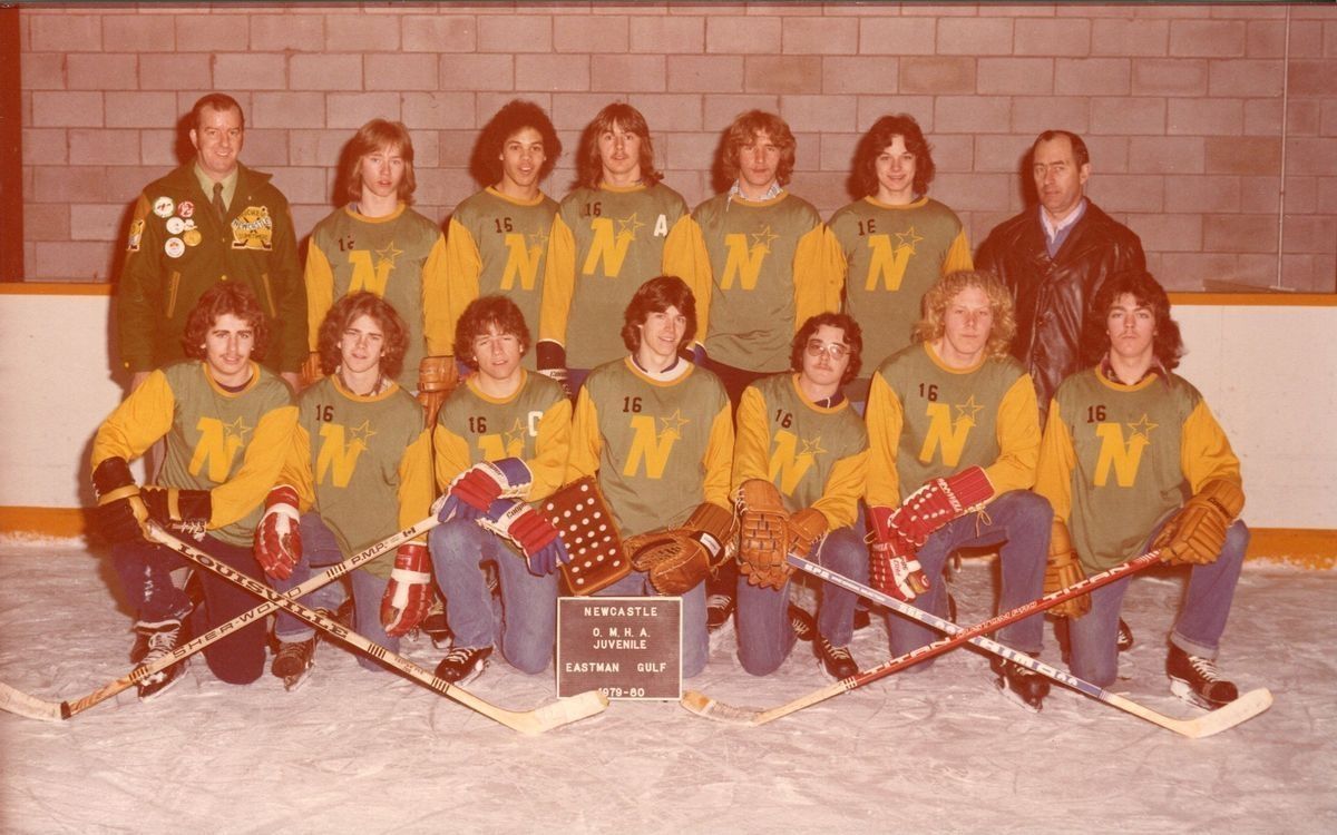 Newcastle OMHA Juveniles team photo.: A colour photograph of twelve young men and two coaches in an arena on the ice. The boys are wearing matching green shirts with yellow sleeves and trimming. the jerseys have a yellow N on the with a star on the N's right side, there also seems to be a black number sixteen on most of the shirts. The boys are also wearing blue jeans and hockey gloves in varying colours. The coach on boys' right side is wearing a dark green jacket with many different patches, and coach on their left side is wearing a black leather jacket under a blue and white collared shirt. In the front row, the first and third as well as the fifth and seventh boys are crossing their sticks. In front of the boys is a black plaque with a white outline that reads 'Newcastle OMHA Juvenile Eastman Gulf 1979-80'. The photograph's colours have faded a bit.