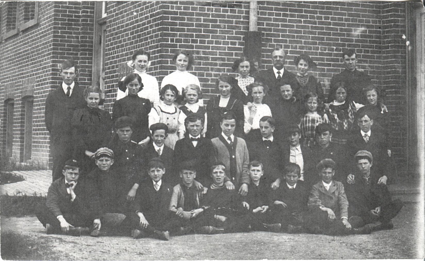 Students at Newcastle High School Circa 1900s: The image shows a group of children and adults posing in multiple rows, dressed in early 20th-century attire, with a brick building as the backdrop. The children are divided into two groups. The boys are sitting crouching, while the girls stand behind them. while the few visible girls stand in the middle. The adults are standing on either side of the group, with another standing closer to the right than the middle.