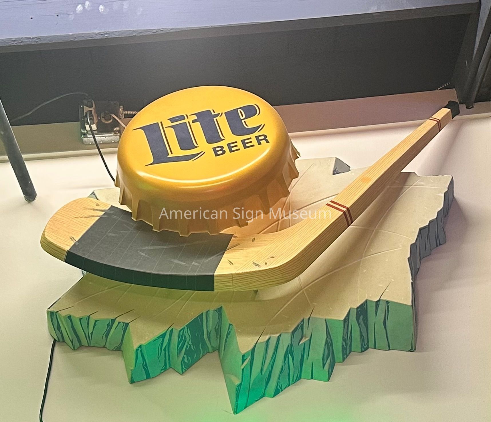 This sign features layered 3D forms of an illuminated lite beer bottle cap over a hockey stick on a slab of ice