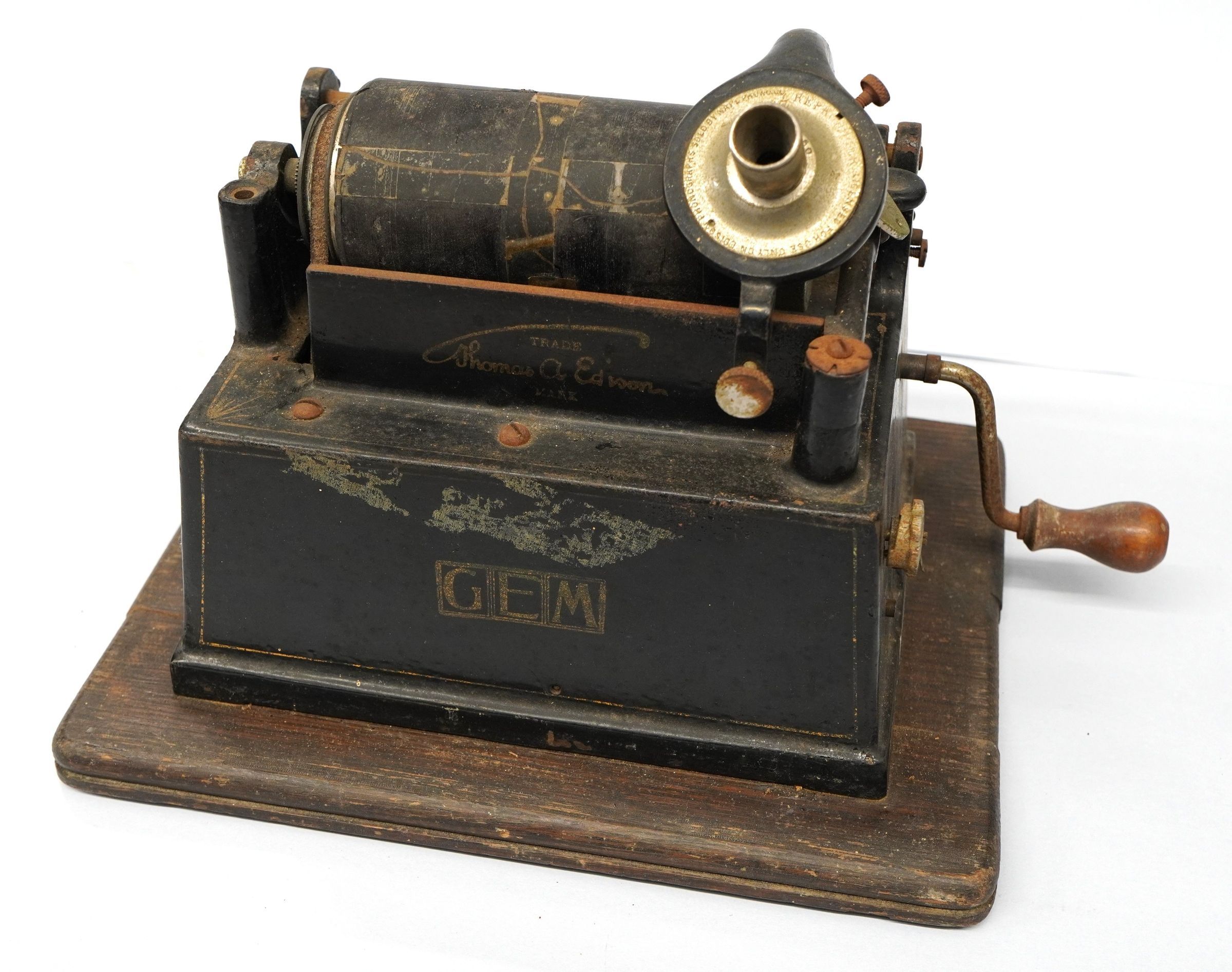 The image depicts an antique phonograph with a wooden base, featuring a black metal body that bears the trademark of Thomas A. Edison, and the word "GEM" on its side, along with visible signs of wear and age, such as scratches and faded paint; it has a prominent crank handle on the side, a cylindrical component on top, and intricate mechanical parts which together showcase the ingenuity and craftsmanship of early sound recording technology.
