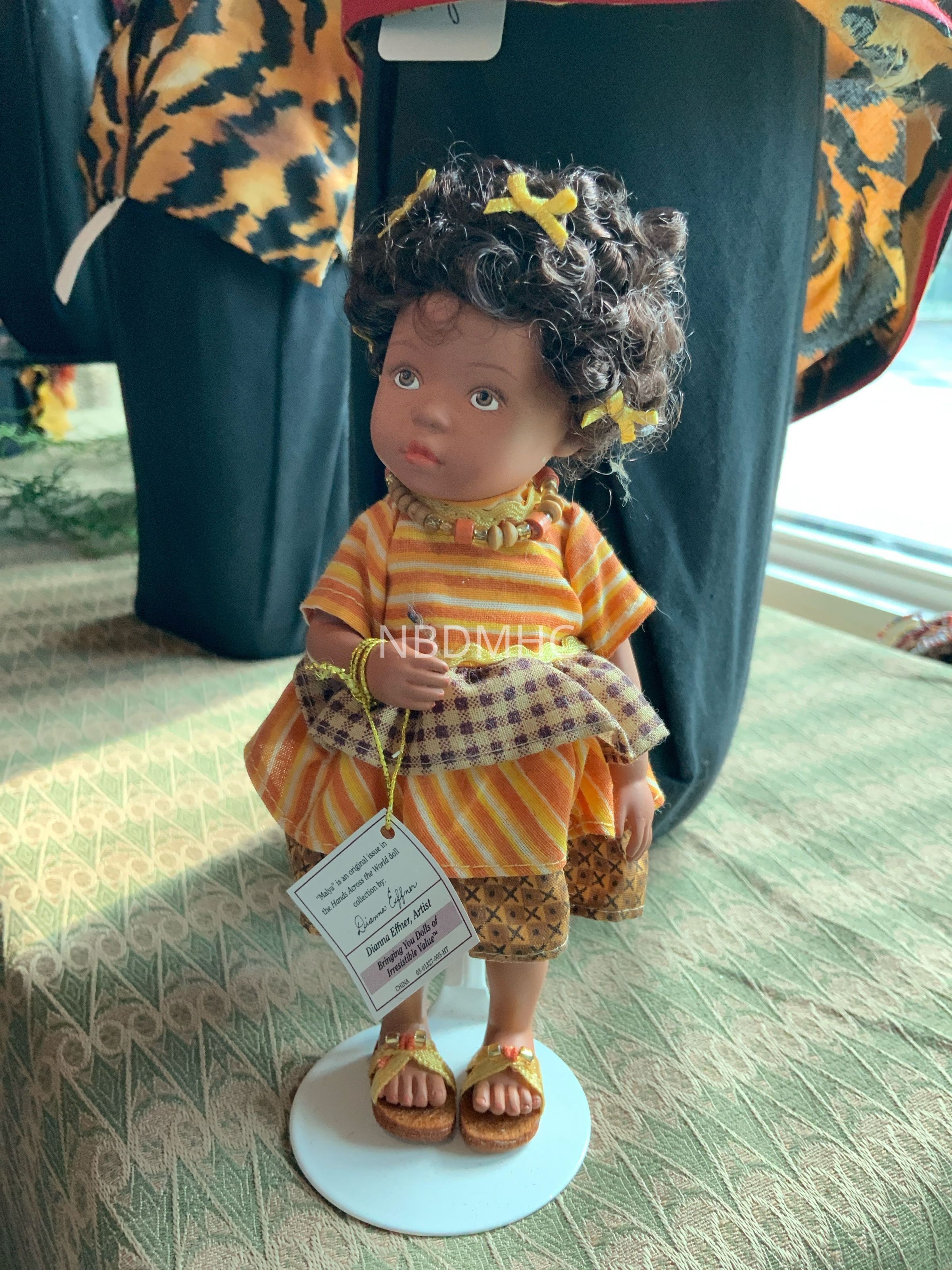 Malya: Hands Across the World Figurines: The image depicts a handcrafted doll standing on a circular white stand, adorned in a vibrant orange and yellow striped dress with intricate patterns at the hem, and accented with a checkered scarf; the doll features curly hair embellished with small yellow bows, and wears sandals that match the ensemble, effectively capturing a charming artisan aesthetic, while a small tag hangs from its wrist, providing details related to the artisan's work, all set against a textured fabric background that complements the overall visual harmony of the scene.