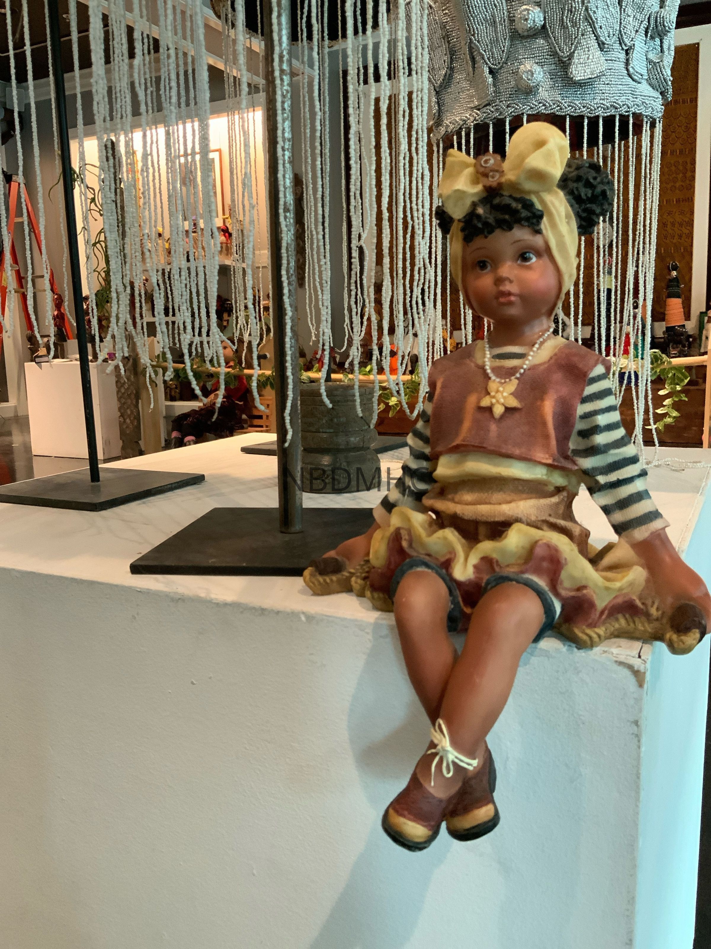 A Girl and her jump rope: The image shows a decorative figurine of a young girl seated on a white pedestal, featured in a museum setting. The figurine is adorned in a vibrant outfit with striped sleeves, a layered skirt with gold and burgundy tones, distinctive brown shoes, and a yellow headband with decorative elements.
