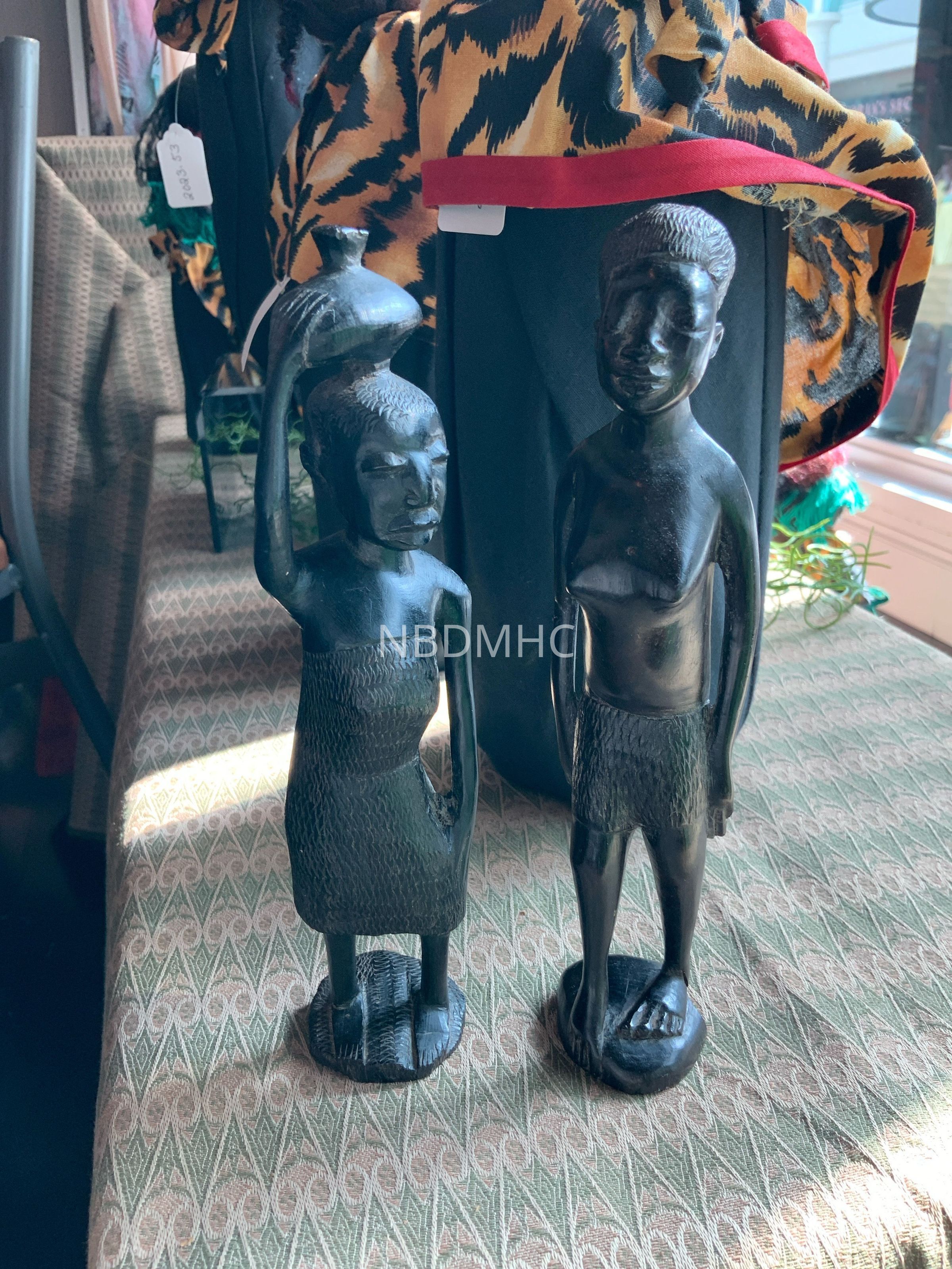 The image depicts two intricately carved wooden sculptures placed on a patterned surface. The sculptures appear to represent human figures, each with distinct postures and attire. The figure on the left holds a jug on its head, suggesting a scene perhaps from daily life or cultural significance, while both are adorned with detailed carvings to represent clothing. Behind them, brightly patterned fabric with a striking yellow, black, and red design drapes over a black backdrop, adding a colorful contrast to the darker tones of the wooden