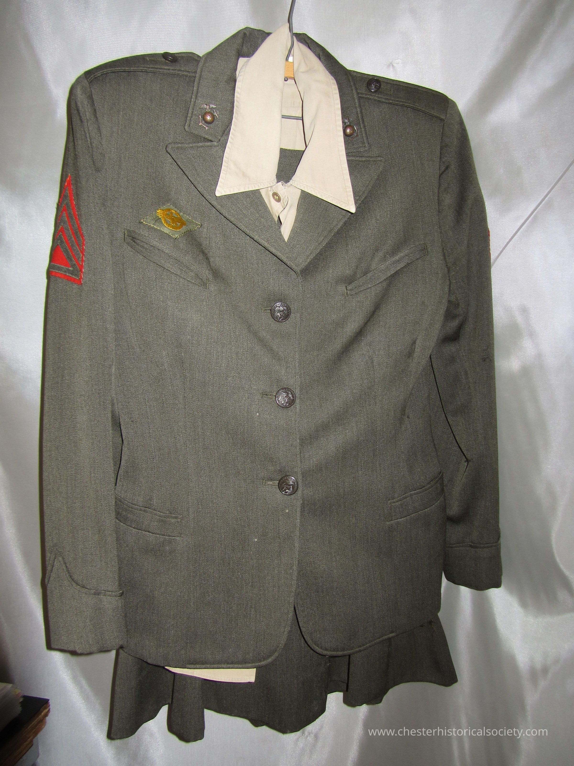 Betty Murray's WWII Marine Uniform: The image depicts Betty Murray's WWII Marine Uniform consisting of a dark green buttoned jacket with a beige shirt underneath, decorated with insignia on the shoulders and chest, and a red chevron patch on the sleeve, all hung on a hanger against a plain background.