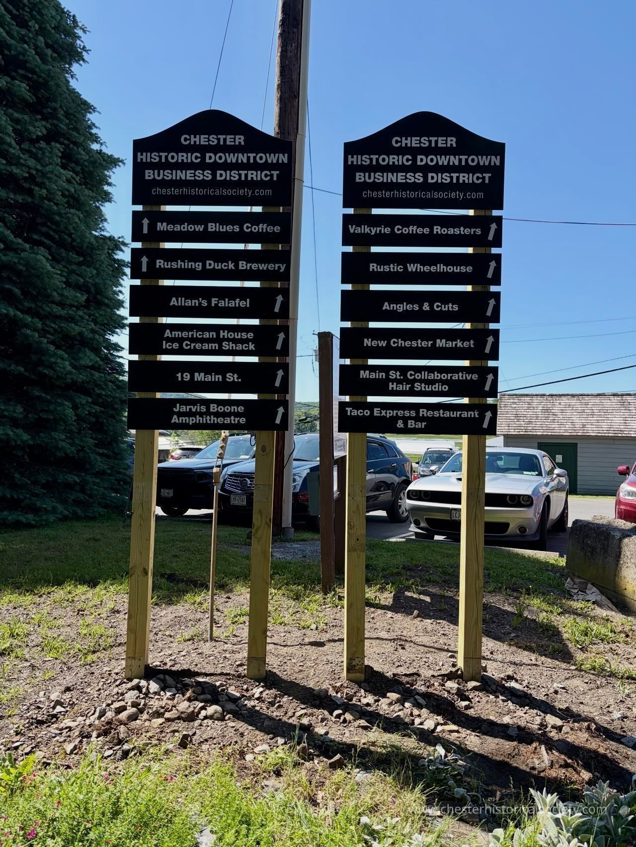 Chester Historic Downtown Business District Trailside Directory: The image depicts two signboards situated on a grassy area, each supported by wooden posts, with the top of each sign reading "CHESTER HISTORIC DOWNTOWN BUSINESS DISTRICT" and providing a website link for the Chester Historical Society. Both signs list various businesses located in the area, with each business name followed by an upward-pointing arrow, suggesting their direction. On the left sign, businesses listed include Meadow Blues Coffee, Rushing Duck Brewery, Allan's Falafel, American House Ice Cream Shack, 19 Main St., and Jarvis Boone Amphitheatre. The right sign lists Valkyrie Coffee Roasters, Rustic Wheelhouse, Angles & Cuts, New Chester Market, Main St. Collaborative Hair Studio, and Taco Express Restaurant & Bar. Behind the signs, several parked cars are visible along with a vibrant green tree to the left.