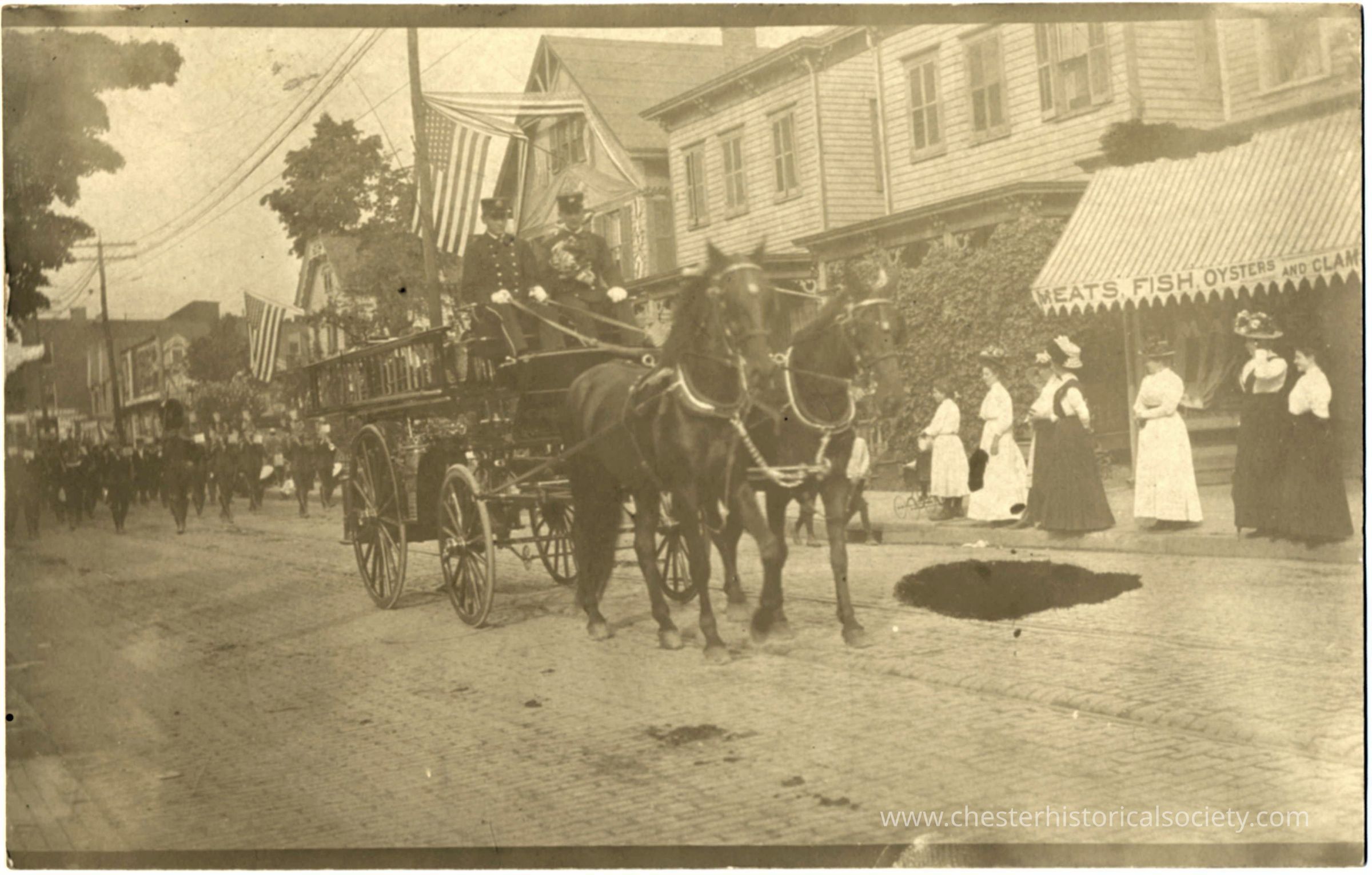 Firemen parading: This monochrome photo postcard a bustling early 20th-century street scene adorned with American flags during a parade, featuring a horse-drawn fire engine , driven by uniformed Chester Firemen. A crowd of onlookers, including women in long dresses and men in suits, lines the cobblestone street, punctuated by a busy storefront.