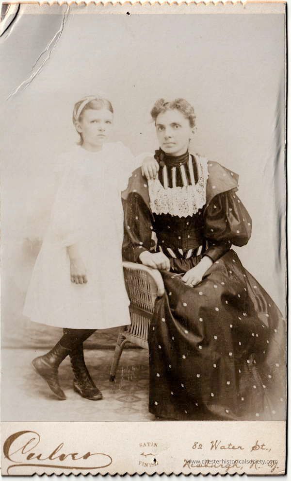 Ms. Charles W. Kerner and daughter Mildred: This vintage black-and-white Cabinet Card photograph, taken by Culver at 82 Water Street, Newburgh, N.Y. features a posed portrait of Ms. Charles W. Kerner and her daughter Mildred. Mrs. Kerner, seated in a wicker chair with an attentive gaze directly at the camera, is attired in a dark dress adorned with a decorative lace collar and ruffled sleeves. She supports the standing girl, dressed in a simple white gown, who looks slightly to the side with a serene expression.