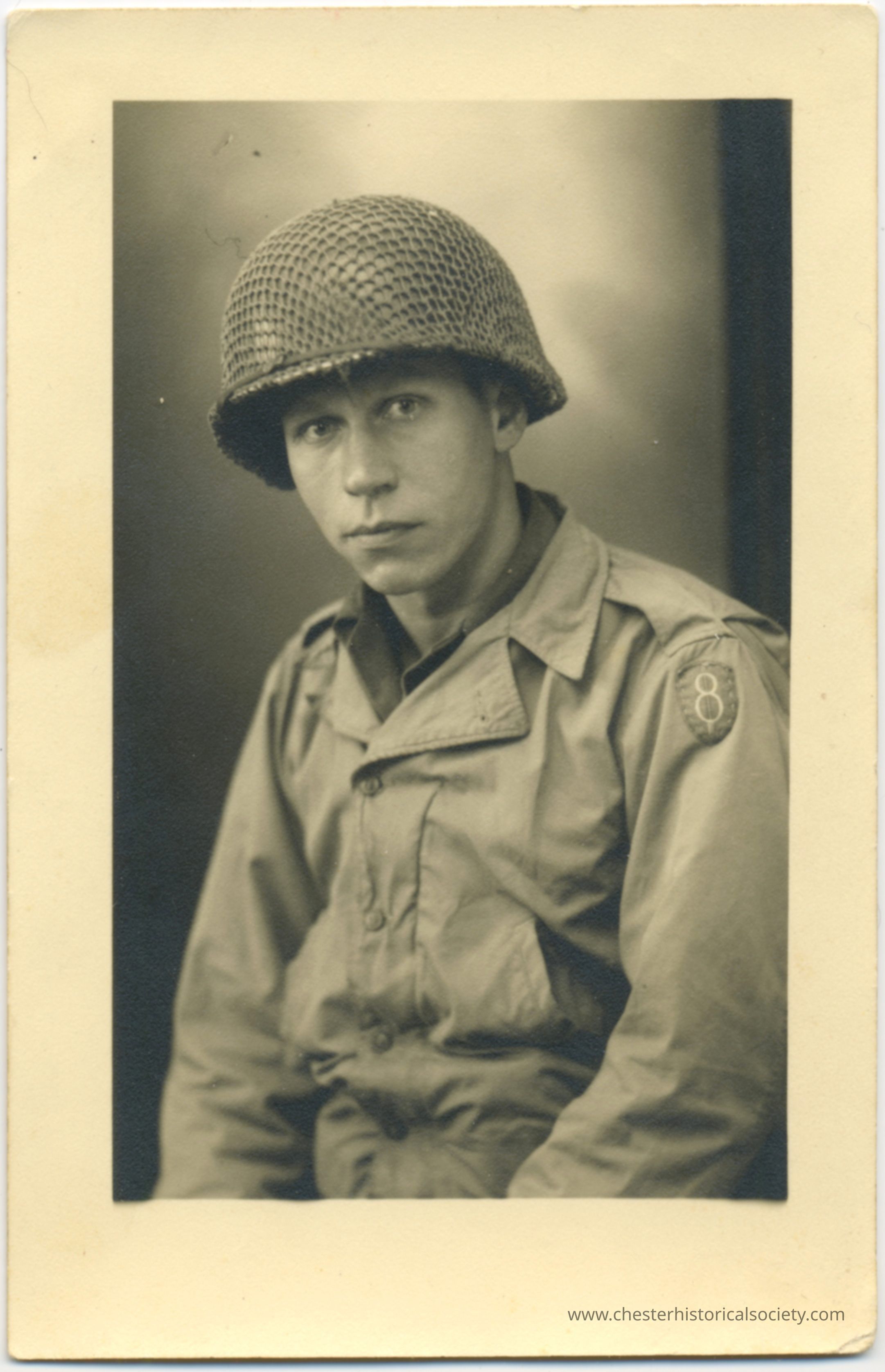 Uncle Alex Flusak: This monochrome photo postcard depicts a young man, Alex Fleesak, dressed in a World War II U.S. Army uniform, staring intently at the camera. He is wearing helmet with a camouflage net and a 8th Infantry insignia visible on his sleeve. His uniform appears worn and rugged, typical of a soldier engaged in active duty. The photograph, monochrome and with a soft focus, captures a moment that seems both personal and intense, emphasizing the seriousness and youth of this soldier.