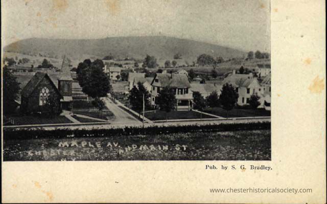 Maple Av. and Main St., Chester, N. Y.: This monochrome photo postcard depicts a vintage photograph of an intersection labeled "Maple Av. and Main St.," showcasing several residential buildings, St. Paul's Episcopal Church, and lush surroundings under a clear sky, with soft rolling hills in the background, capturing the essence of a serene, small town. Published by S. G. Bradley.