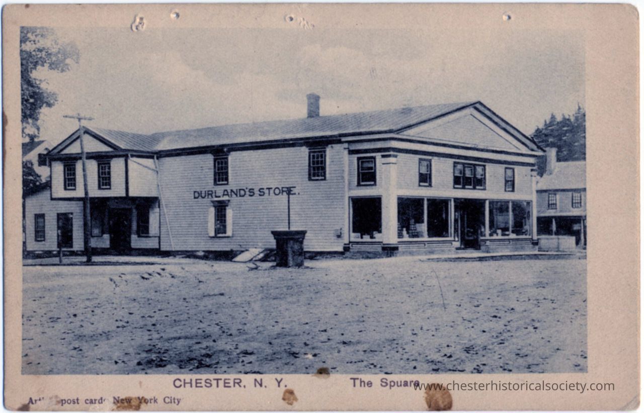 The Square - Durland's Store, Chester, N. Y.: This monochrome photo postcard depicts Durland's Store, located in Chester, N.Y., at the inspection of Academy Ave. and Main St. known as "The Square." The photo showcases a large, two-storied structure with several visible entrances and a chimney, indicating that it functioned both as a store and warehouse. The building is clad in weathered siding and features a multi-windowed facade. In the foreground, a dirt road and a walkway lead to the storefront, and a standalone water trough, in the center of this intersection with a kerosine streetlamp mounted in it.