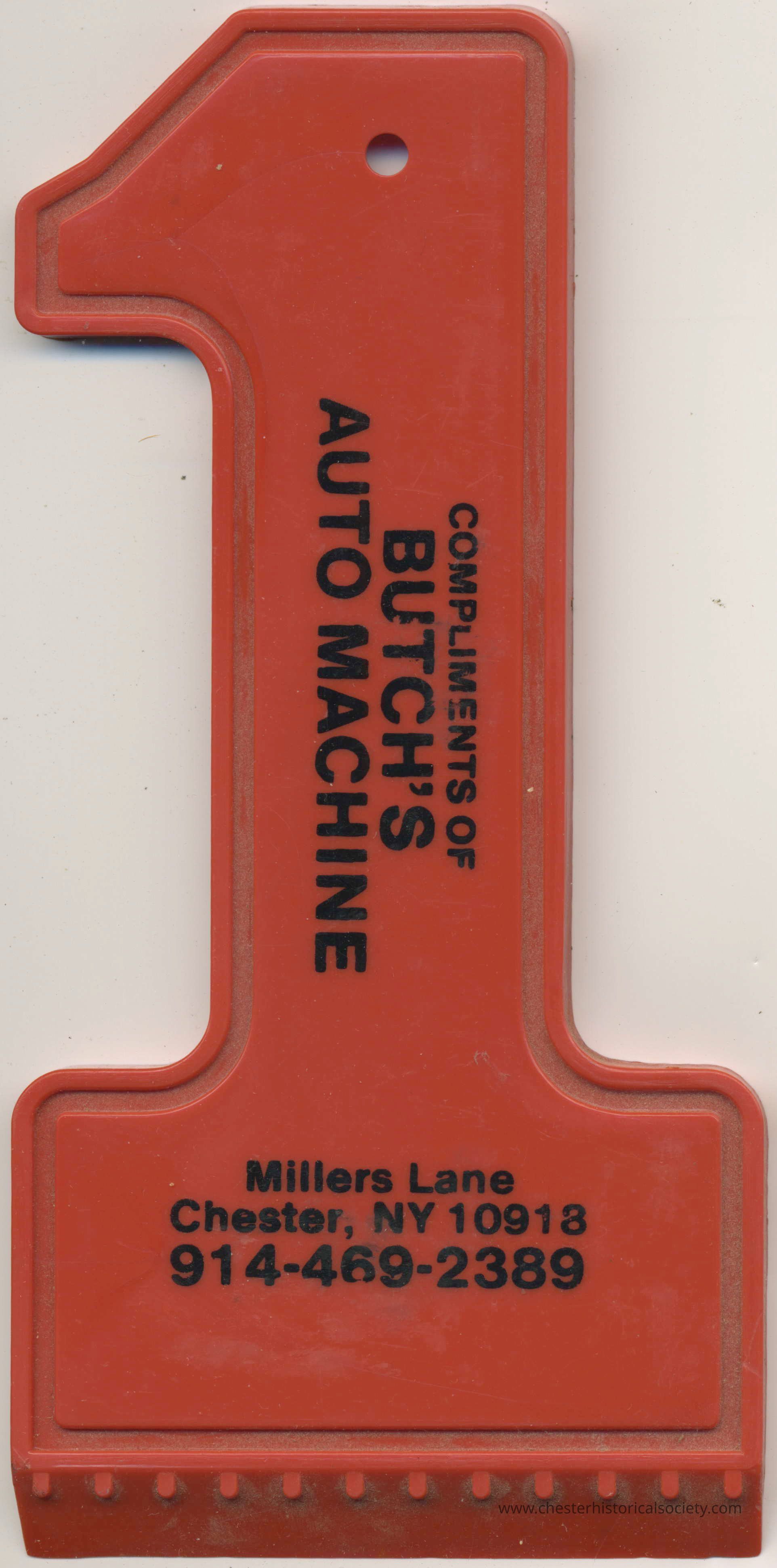 Butch's Auto Machine Ice Scraper recto: The image depicts a red, number 1-shaped promotional ice scraper featuring black text that reads, "COMPLIMENTS OF BUTCH'S AUTO MACHINE," with additional contact information provided below, which includes "Millers Lane, Chester, NY 10918" and a phone number "914-469-2389"; this plastic tool appears to have a hole for hanging at the top and a serrated edge for scraping ice at the bottom, implying its intended use as a practical giveaway item from a local business.