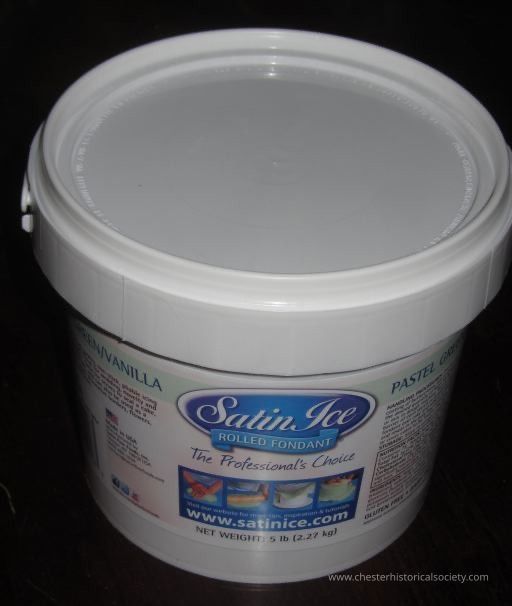 Pastel Green/Vanilla Rolled Fondant 5 lb Container: The image shows a container of Satin Ice Rolled Fondant, prominently labeled as "The Professional's Choice," featuring a secure, snap-on lid in a neutral light gray color; the packaging details its net weight as 5 pounds (2.27 kg), and highlights the item’s intended use in cake decoration and design, showcasing sample images of the product in vibrant, pastel colors on cakes, while providing the website www.satinice.com for further product information.