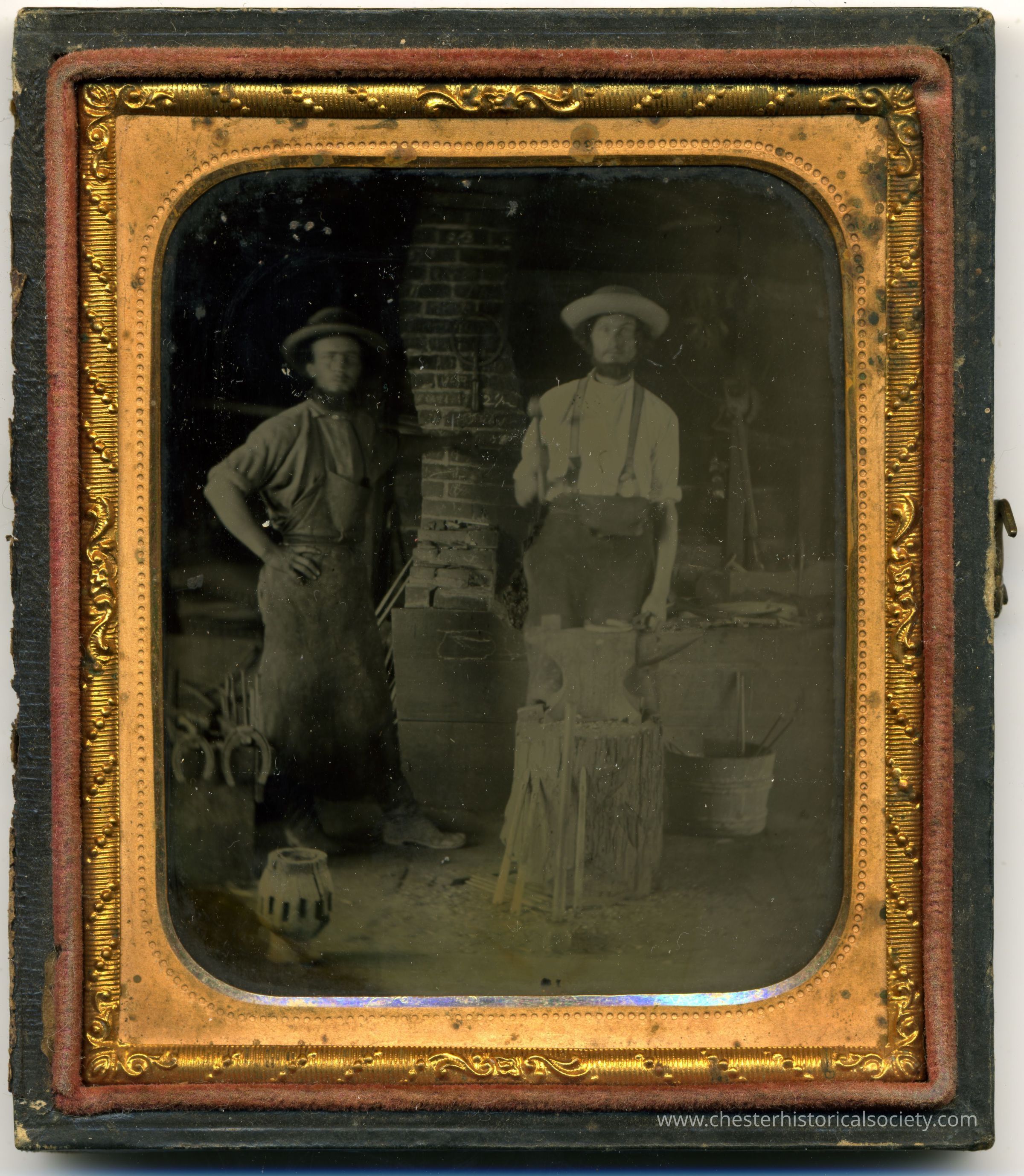 Two blacksmiths: The image depicts two men standing in a rustic workshop-like setting, featuring tools, buckets, and a brick chimney. The man on the left stands casually with one hand in his pocket, wearing a rounded hat and dungarees, while the man on the right, wearing a wide-brimmed hat, suspenders, and a rolled-up shirt, stands more squarely. They are framed by an ornate, golden, embossed border. The case is missing its cover.