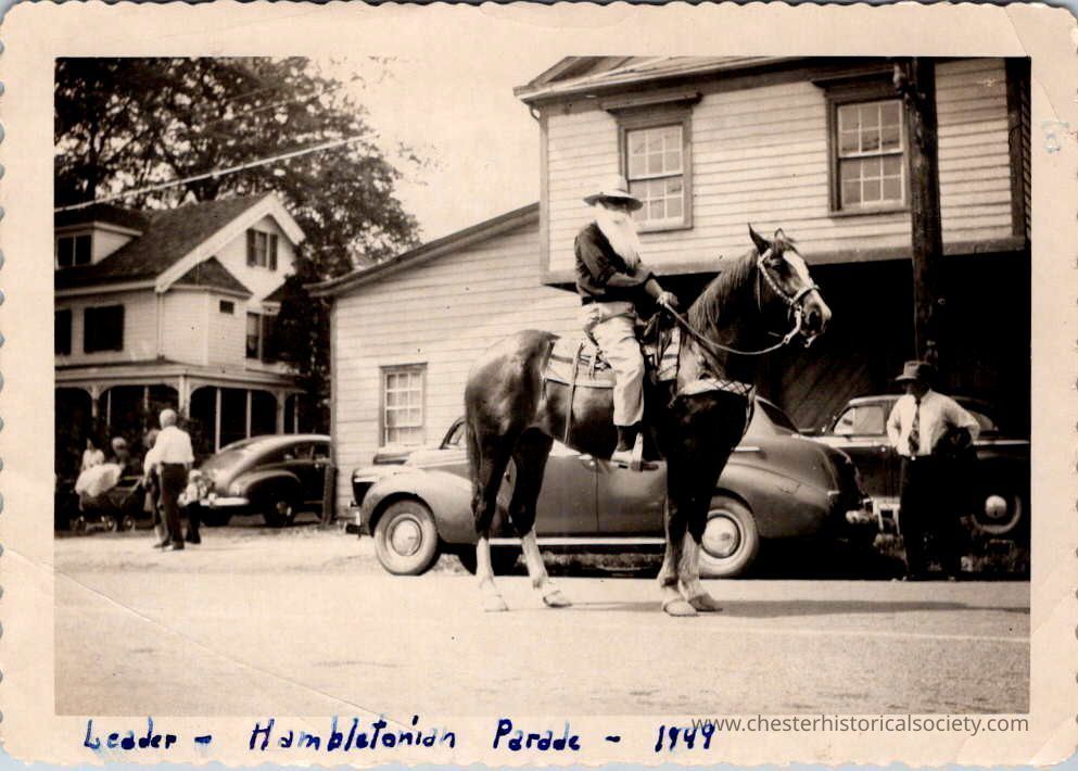 Leader - Hambletonian Parade - 1949: The black-and-white photograph depicts a scene from the 1949 Hambletonian Parade, showing William R. (Bill) Conklin on horseback dressed as William Rysdyk, leading the procession down Academy Avenue in the Village of Chester, lined with older model cars and spectators.