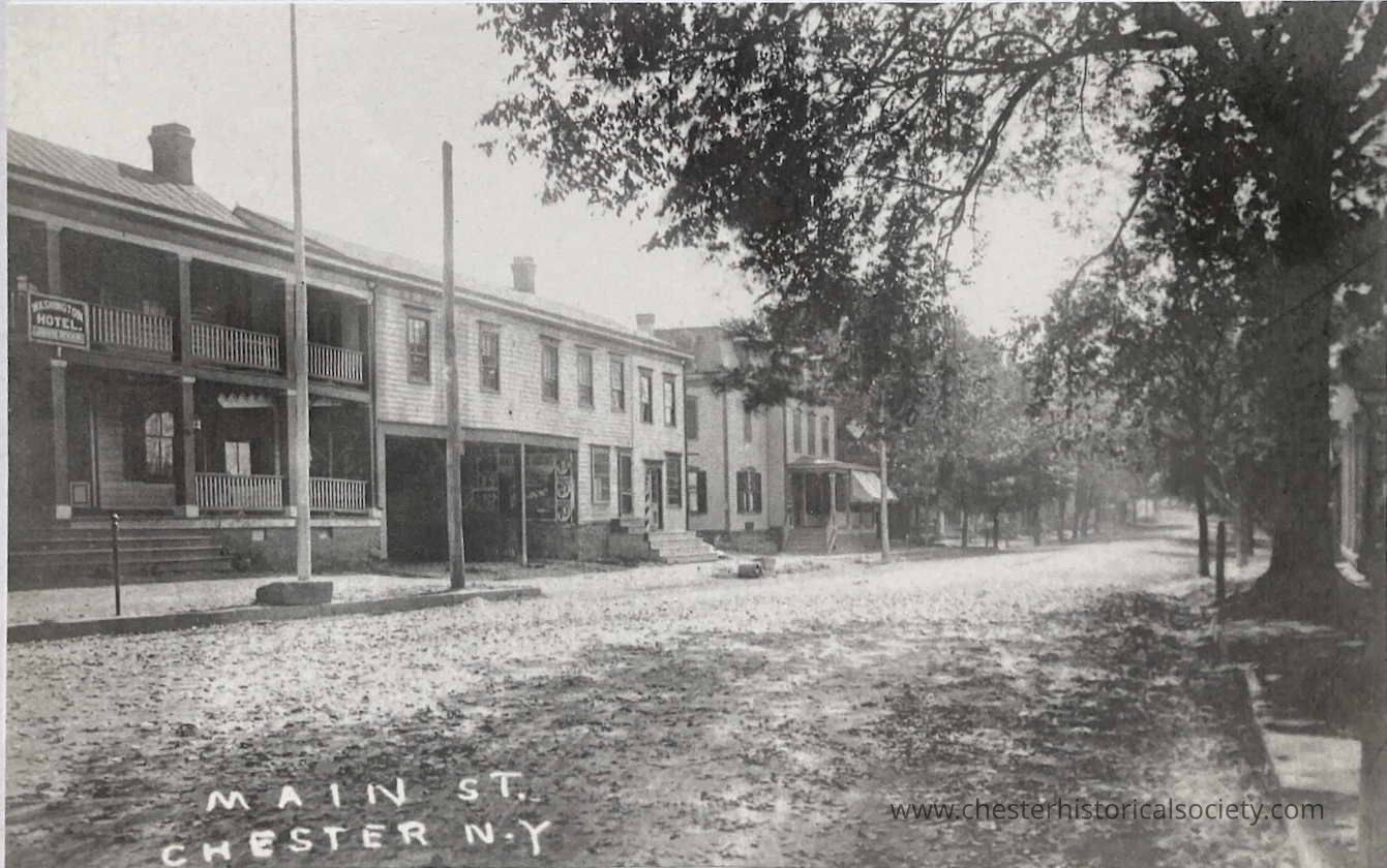 Image of a post card titled Main St., Chester, N.Y. that clearly displays the Washington Hotel sign: The image captures an undated view of Main Street in Chester, New York, featuring a row of two-story buildings with balconies, notably the Washington Hotel hotel, bordered by dirt roads and mature trees. Signs and stairs adorn the wooden structures while shadows and sunlight play across the scene, reflecting the architecture and character of 'Uptown' Chester, Orange County, New York in the turn-of-the-century period.