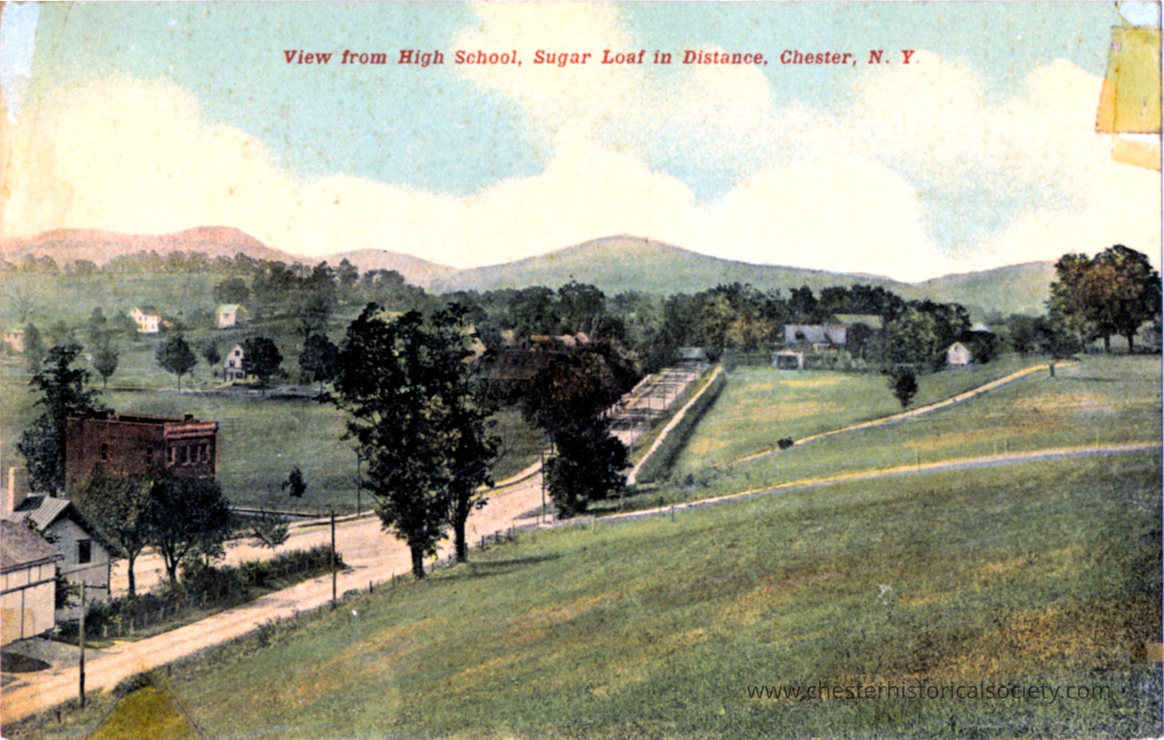 View from High School, Sugar Loaf in distance, Chester, N. Y.: This colorized photo postcard presents a tranquil panoramic view from a high school in Chester, New York, depicting lush green fields and scattered buildings under a vast sky, with the shadowy outline of Sugar Loaf Mountain visible in the distance. The image captures the essence of a serene, pastoral landscape, marked by its rural charm and the simplicity of early 20th-century American life.