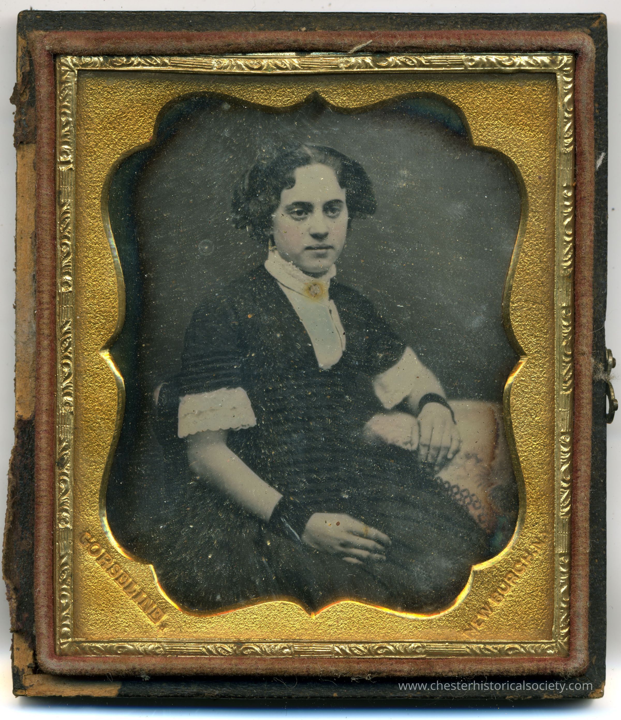 Photograph of a young lady seated: The image depicts a vintage portrait of a woman with dark hair and a serious expression, dressed in a black dress with white cuffs and a white collar, seated with her hands relaxed. The photograph is encased in an ornate gold-colored frame with embossed detailing and darkened edges, suggestive of considerable age, contained within a worn leather folding case whose lid is missing.