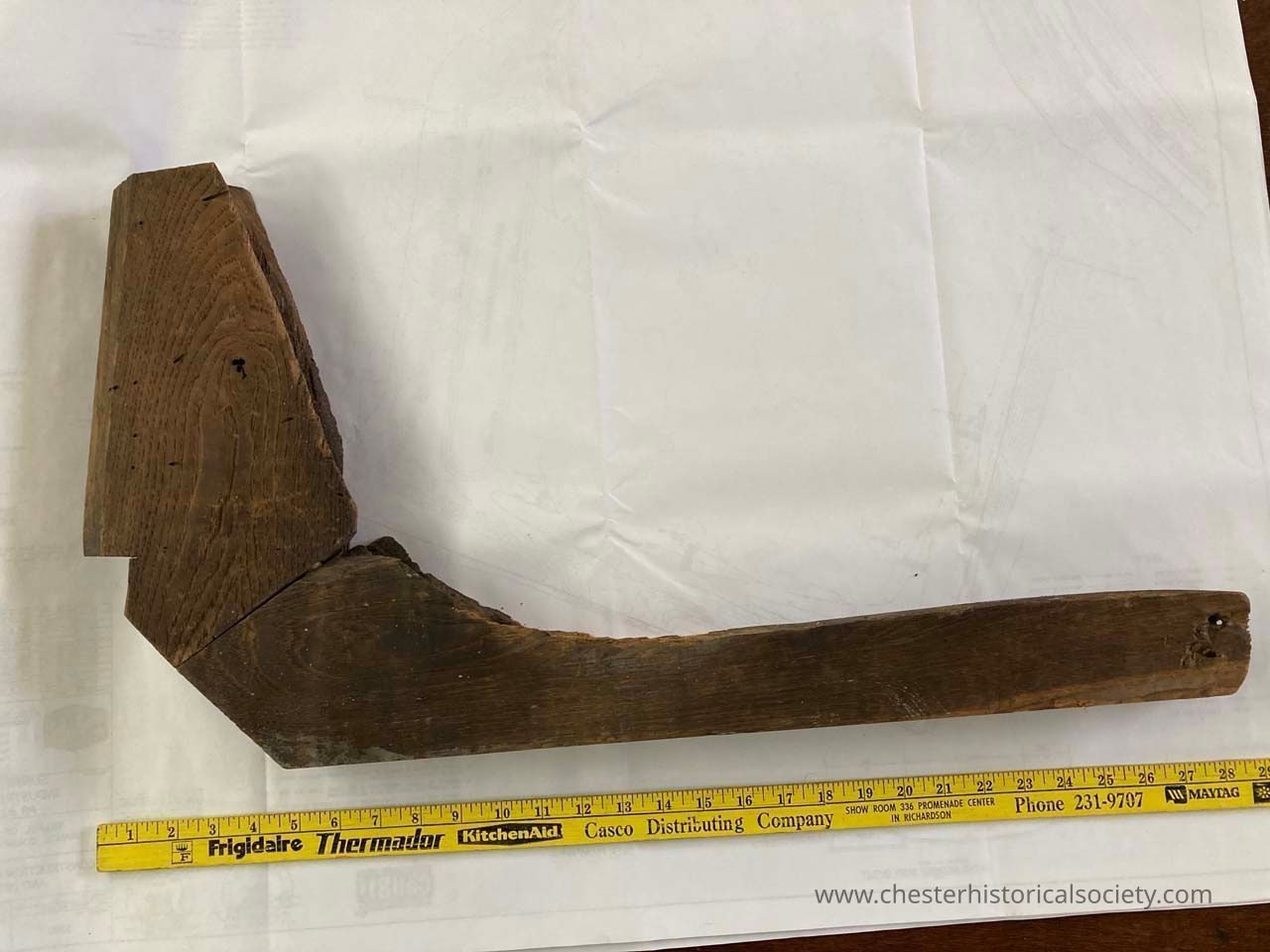 Section of Lady's Waiting Bench Corner Frame - left side: The image features a rustic, L-shaped piece of wooden furniture or structural element resting on a white surface, with visible grain patterns and slight weathering. Along the bottom edge, there is a yellow stick, providing a sense of scale. The wood is darker at the thicker, vertical section, which angles sharply into the longer, horizontal arm, appearing slightly rough-hewn and aged. The entire setup is viewed from above, showing the texture and natural imperfections of the assembly.