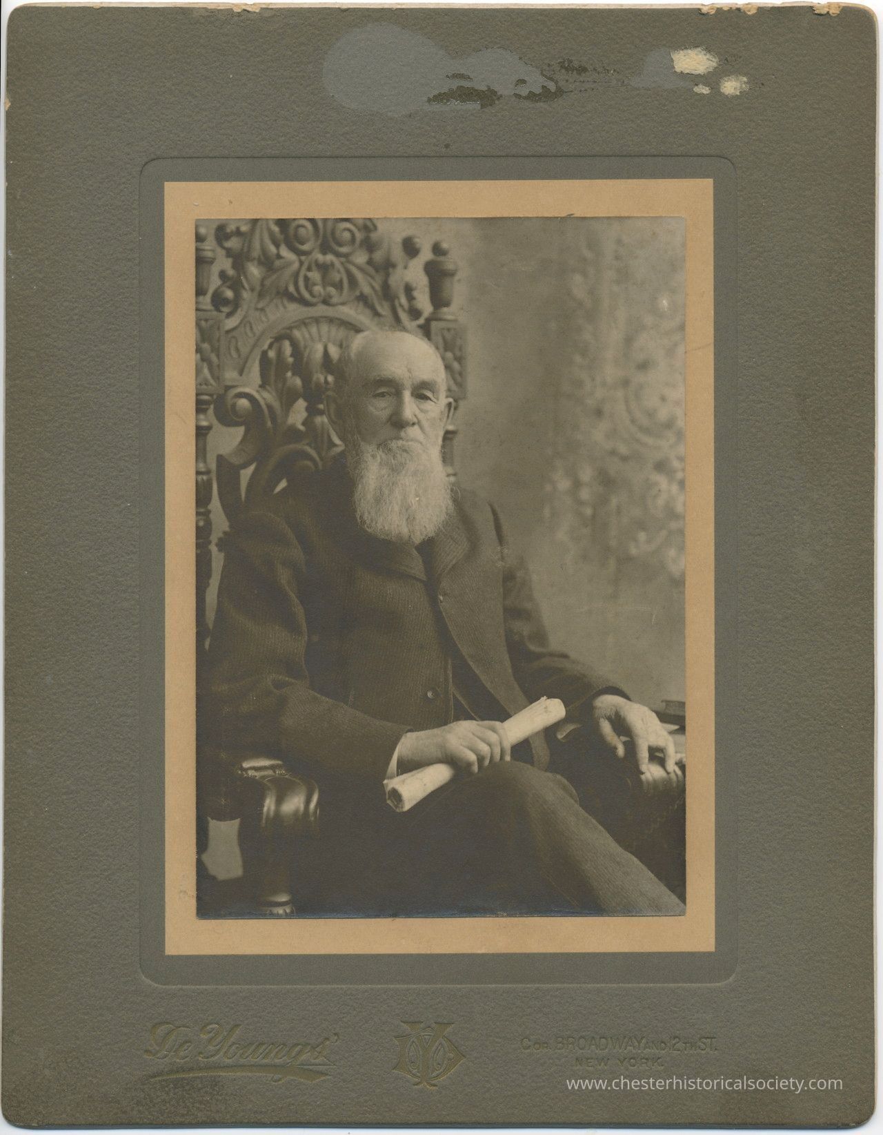 Jonathan Hudson Board: The sepia-toned photograph depicts Jonathan Hudson Board as an elderly man with a long white beard seated in an ornately carved wooden chair, holding a rolled document in his right hand, suggesting a formal or ceremonial setting; the man's attire consists of a dark suit, and he appears calm and composed, embodying the characteristics typical of a distinguished figure of his era, while the elaborate chair and patterned backdrop suggest a studio setting.