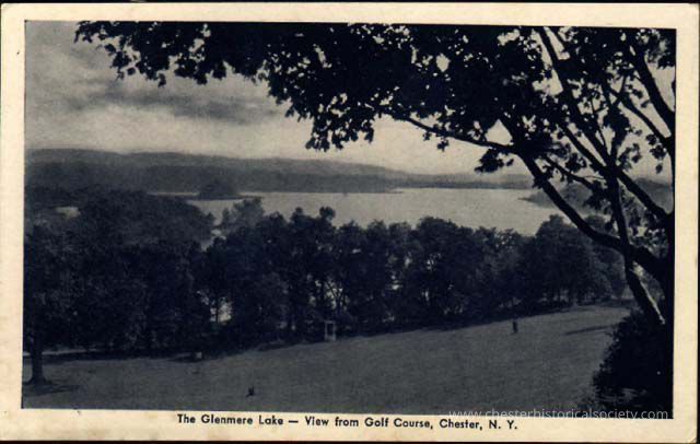 The Glenmere Lake – View from Golf Course, Chester, N.Y: This monochrome photo postcard presents a serene view of Glenmere Lake as seen from a golf course in Chester, New York, capturing the expansive water body bordered by lush forests under a cloudy sky, with the foreground showcasing a well-maintained grassy slope dotted with trees, offering a picturesque and tranquil landscape.
