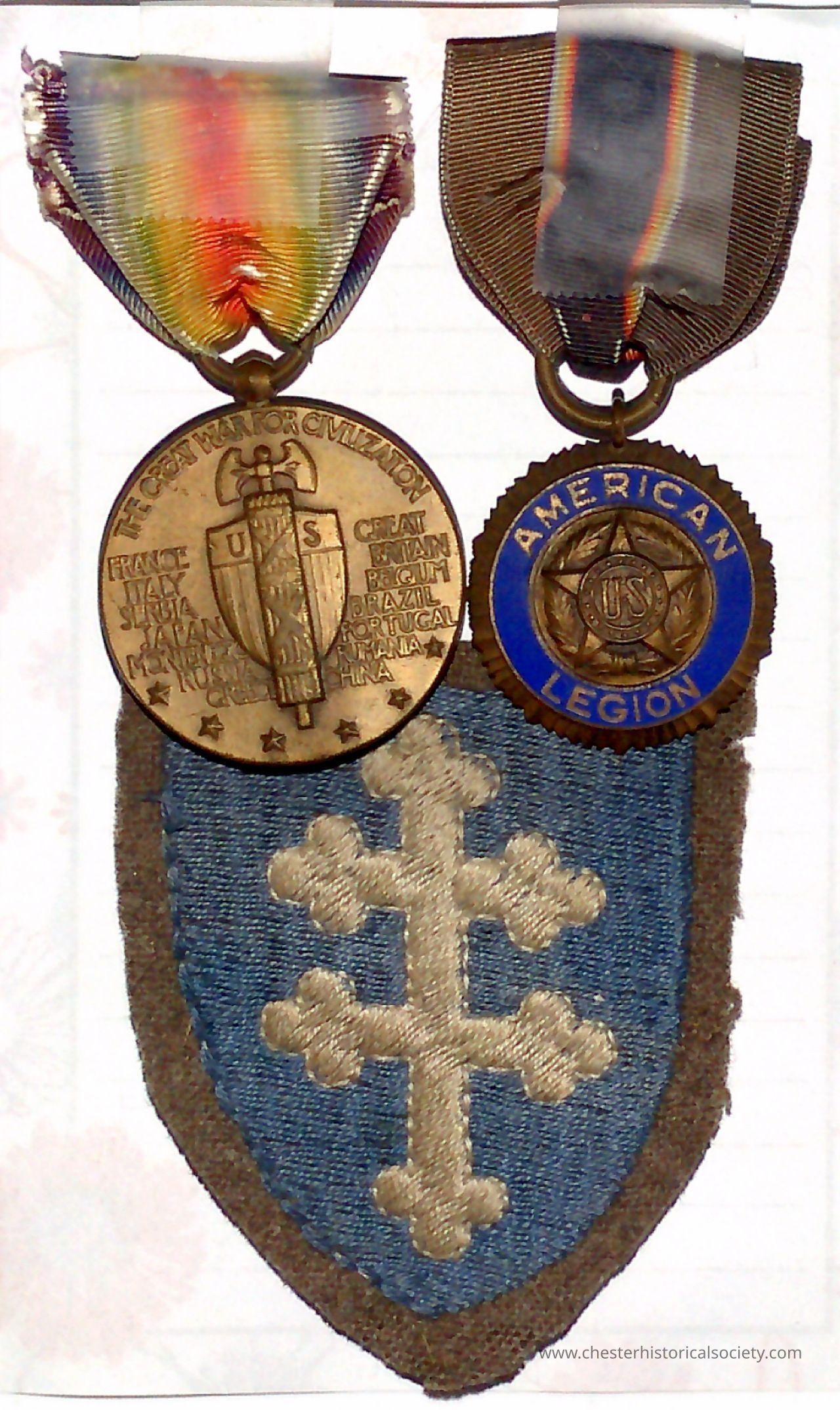 Charles Napp's WWI Victory medal, Amerion Legion Medal & 79th Div. unit patch: The image displays Charles Napp's two medals and an embroidered patch. The left medal features an inscription surrounded by a rainbow ribbon, depicting a shield with an eagle, while listing several countries. The right medal has a dark ribbon and a circular design with the words "American Legion" in blue around its perimeter, with a star and the initials "USS" at the center. Below the medals is a patch shaped like a shield, predominantly blue with a brown border, showcasing a beige emblem of a double-barred cross.