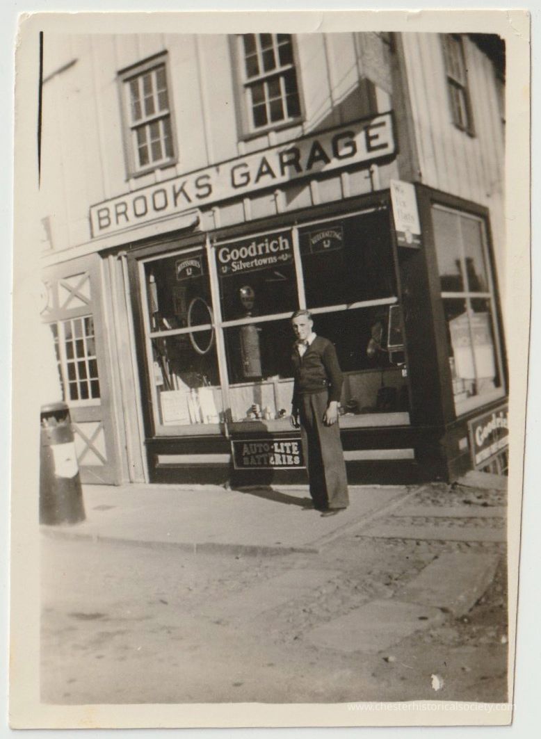 The image depicts a scene outside an establishment named "Brooks Garage," adorned with signage advertising Goodrich Silvertown tires and Auto-Lite batteries. A man stands in front, dressed in early-mid-20th-century period apparel. The façade showcases large windows filled with automotive products, highlighting the garage's function as a point of sale and service for automobile essentials, situated on a street corner neatly paved with cobblestone and slabs.