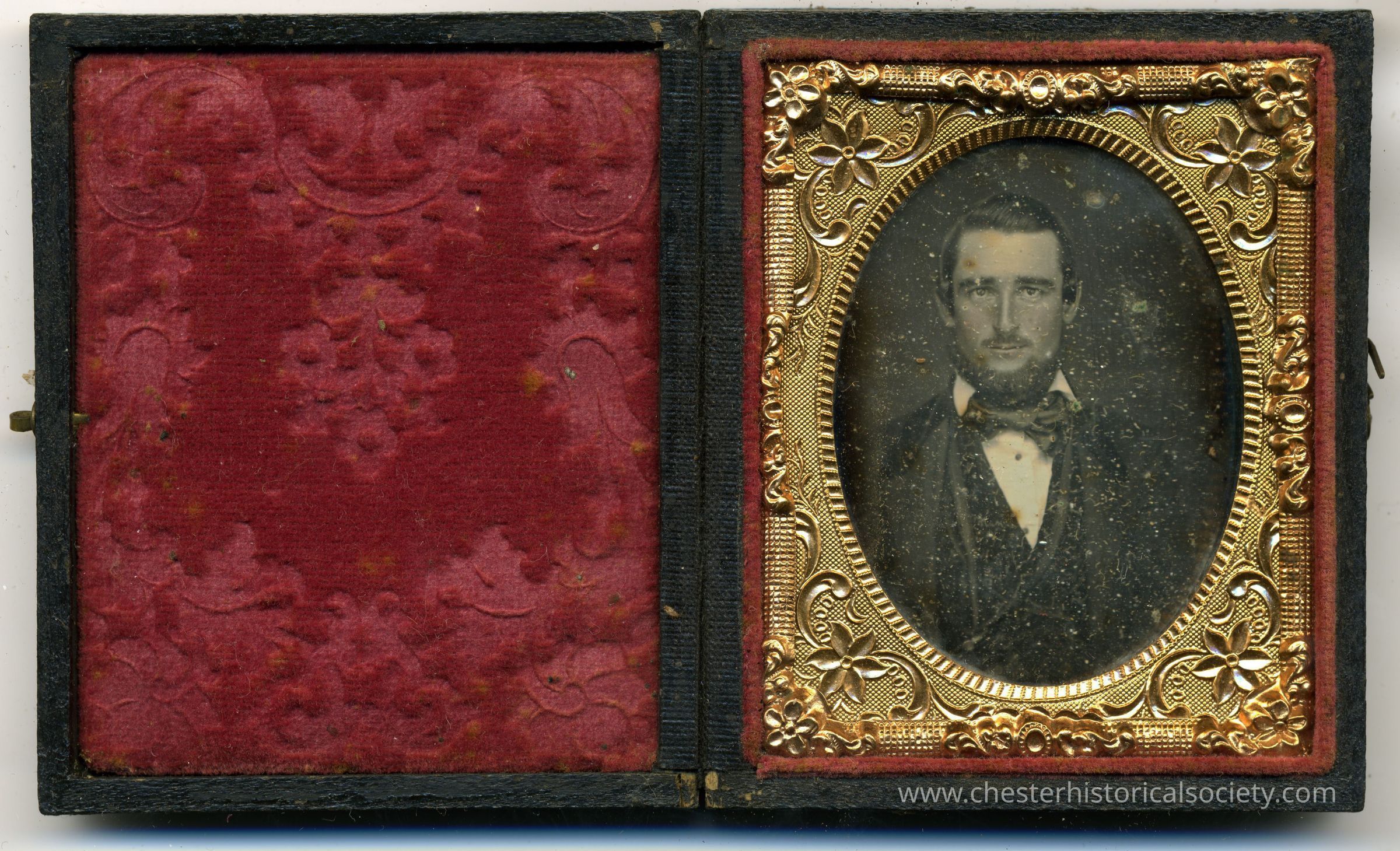 A cased daguerreotype of a bearded gentleman wearing a bow tie.: The image depicts a daguerreotype portrait of a man from the mid-19th century, housed inside an ornate gold frame with floral motifs and enclosed in a red velvet-lined folding case. The man, dressed in a dark suit and bow tie, gazes solemnly forward, his visage rendered in monochrome, highlighting the historic photographic technique. The case's exterior shows wear, suggesting its age and frequent handling over decades, preserving a moment in time with remarkable clarity and detail.