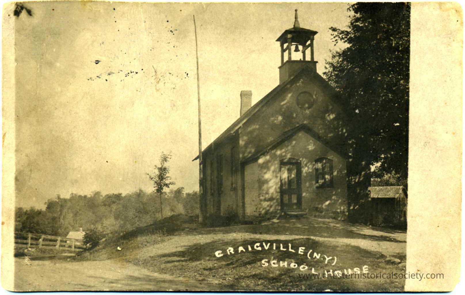 Craigville (N.Y.) School House: This monochrome photo postcard depicts an old schoolhouse in Craigville, NY, featuring a simple, rustic design with a bell tower and large windows, framed by a backdrop of trees and a wooden fence on the left; the building, nestled on a gentle slope, exudes a sense of historical charm, emphasized by the soft light filtering through the surrounding foliage, casting dappled shadows on the ground and structure, while the handwritten label at the bottom of the image marks its identity and location.
