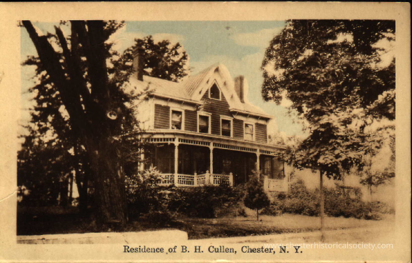 Residence of B. H. Cullen, Chester, N. Y.: This colorized photo postcard depicts the residence of B. H. Cullen, located on High Street, Chester, New York. The house is a beautiful, two-story Victorian structure with a distinctive steeply-pitched roof and decorative gables. A wrap-around porch, adorned with ornate railings, extends around the visible front and side of the home. The house is nestled amidst mature trees and lush greenery, suggesting a serene, rural setting. The photo captures a bygone era
