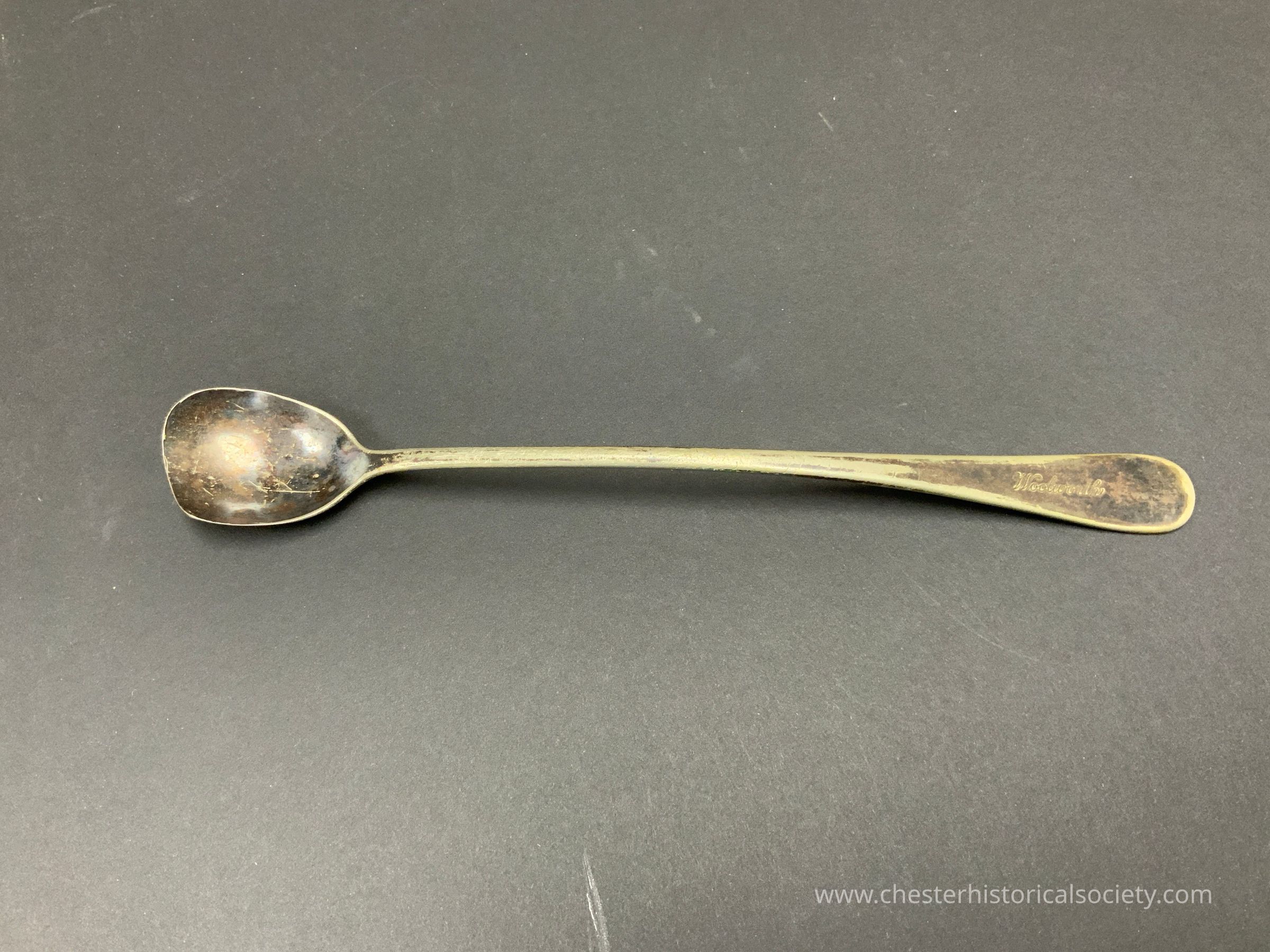 A tarnished silver spoon showing signs of wear and age, with scratches and discoloration predominantly visible on its bowl and along the handle. There is an inscription etched into the handle, Woolworth. The long, slender shape of the spoon handle contrasts with the rounded bowl, providing a hint of elegance despite its worn condition. The overall simplicity and the patina speak to its utilitarian history, possibly having served countless meals over the years.