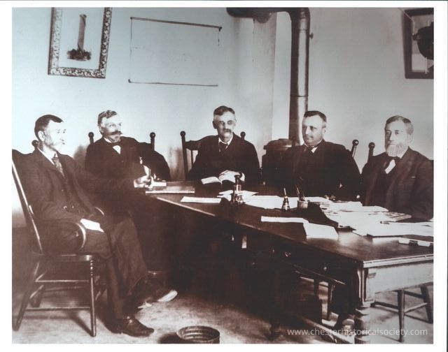 Officers of the first Corporate Village of Chester 1892: From left - Trustee, Thaddeus S. Durland; Treasurer, Charles W. Kerner; President, William A. Lawrence; Trustee, George M. Roe & Trustee, Joseph Durland.
In a sepia-toned historical photograph, five men are seated around a large wooden table in a room, which appears to be a meeting or conference space, characterized by a sense of formality and seriousness. The table is cluttered with various papers and writing materials, indicating preparations for a discussion or decisions on significant matters. The men, dressed in formal suits and ties common to a bygone era, exude an aura of authority, likely suggesting roles of importance and responsibility, perhaps as members of a committee or council. Their expressions range from thoughtful to attentive, with some holding writing instruments, suggesting engagement with the content before them. The room is simply decorated, with a visible framed picture on the wall and a heating stove pipe running into the ceiling corner, hinting at a setting from the late 19th to early 20th century. Simple chairs with high backs surround the table, reinforcing the formal atmosphere. This composition captures a moment in time where decisions and deliberations might have been key