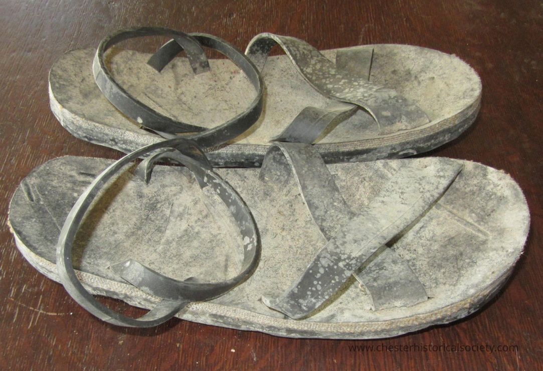 Vietnam style sandals made from tires: The image displays an old, worn-out pair of sandals with dusty, faded surfaces placed on a dark wooden floor, showing signs of heavy usage with spots and discoloration on the straps, and the soles exhibiting visible wear and tear, suggesting a long period of service and exposure to various elements over time, highlighting the robust and enduring nature of these simple footwear items that have probably seen many journeys, reflecting their practicality and resilience despite the apparent rough conditions they have endured.