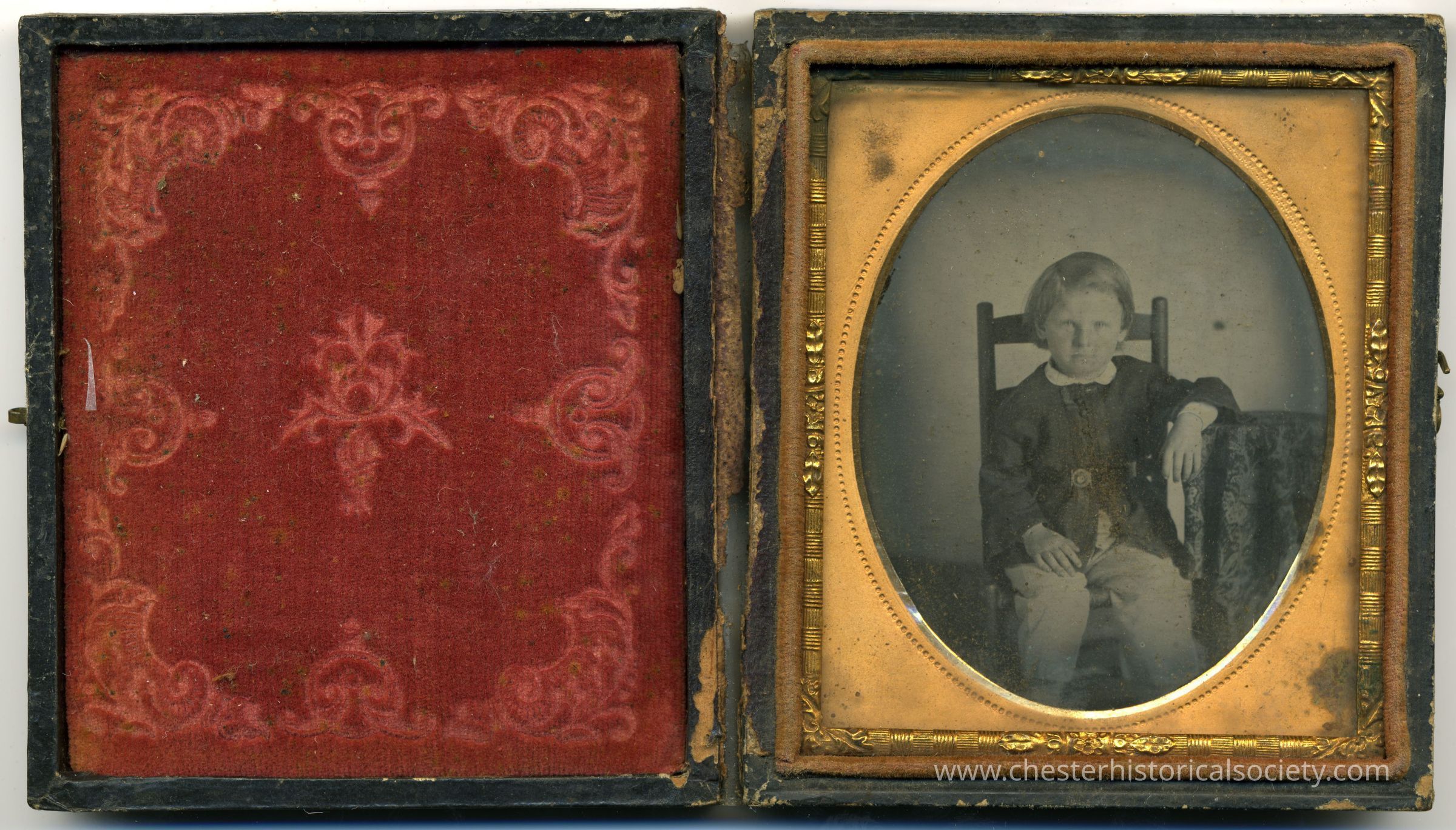 The image displays an old-fashioned hinged photograph case, one side revealing a richly detailed red fabric with ornate designs, and the other side showcasing a sepia-toned daguerreotype portrait of a young child seated in a wooden chair, surrounded by an oval gold-tone frame, capturing a glimpse of the past.