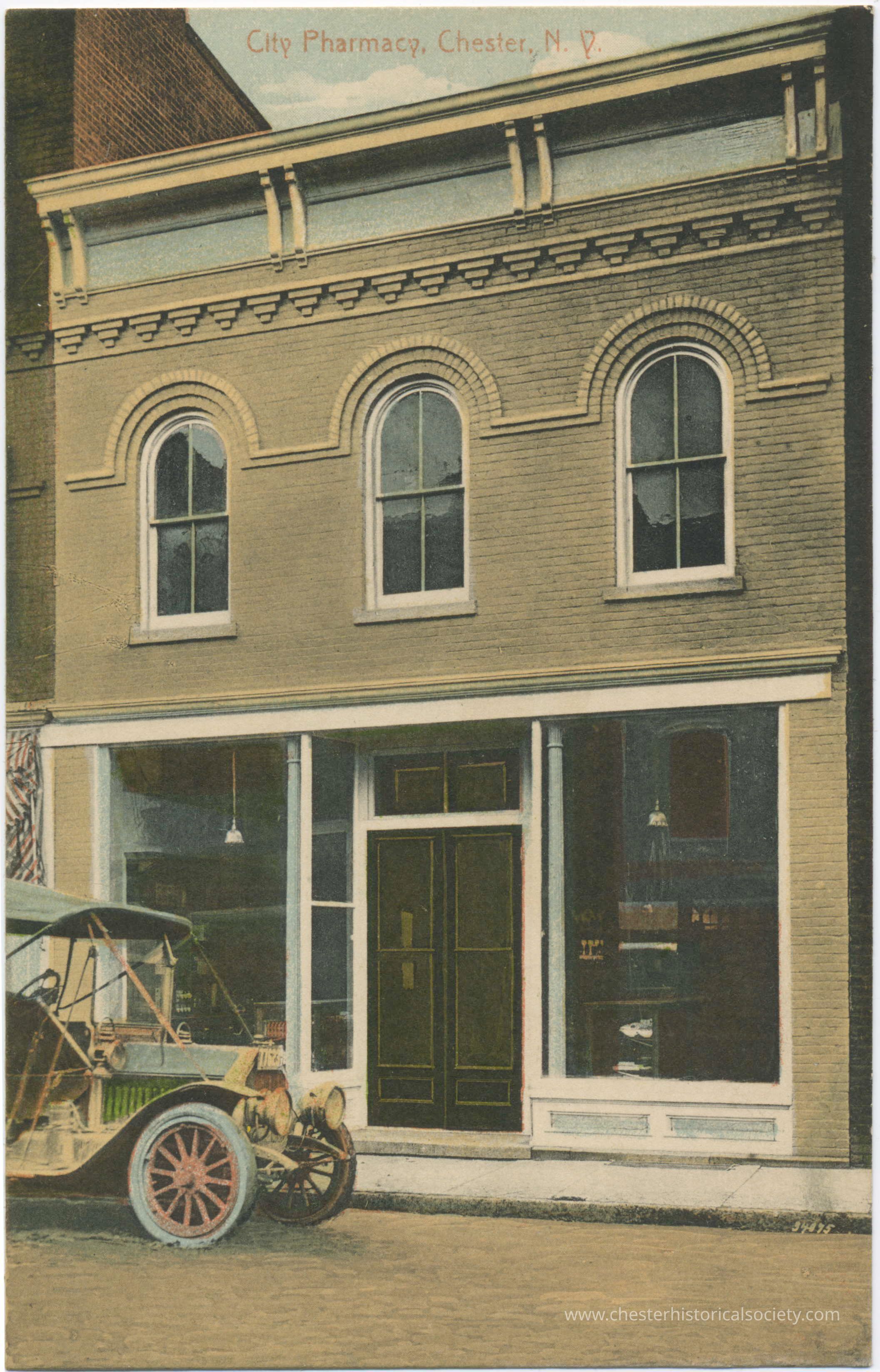 City Pharmacy, Chester, N.Y.: This colorized photo postcard shows City Pharmacy located in downtown Chester, NY, featuring a two-story brick building with decorative detailing and arched windows. In front, an early-model open automobile is parked on the street, indicating a period likely in the early 20th century. The pharmacy's windows display various items, suggesting it is a place offering sundry goods alongside medications—capturing a moment in time when local pharmacies served as crucial community hubs.