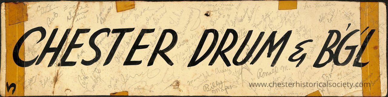 Chester Drum & B'G'L Monroe Parade sign: The image shows a rectangular, vintage-looking paper sign with the bold text "CHESTER DRUM & B'GL" in large, black, capital letters at the center. The background is a yellowed, aged paper. Surrounding the central text are numerous handwritten names and signatures of cor members in a variety of cursive styles, used to memorize the 1950 Memorial Day Parade participants. The edges of the paper are taped with brown tape, showing signs of wear.