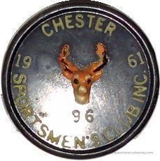 1961 Chester Sportsmen Club pin #96: The image displays a circular plastic badge with a black background featuring a small, central depiction of a deer's head, showcasing detailed antlers in brown and cream colors. Surrounding the deer emblem, the badge is inscribed with text, including "CHESTER SPORTSMEN'S CLUB INC." around the edge, with the year "1961." At the bottom, the number "96."