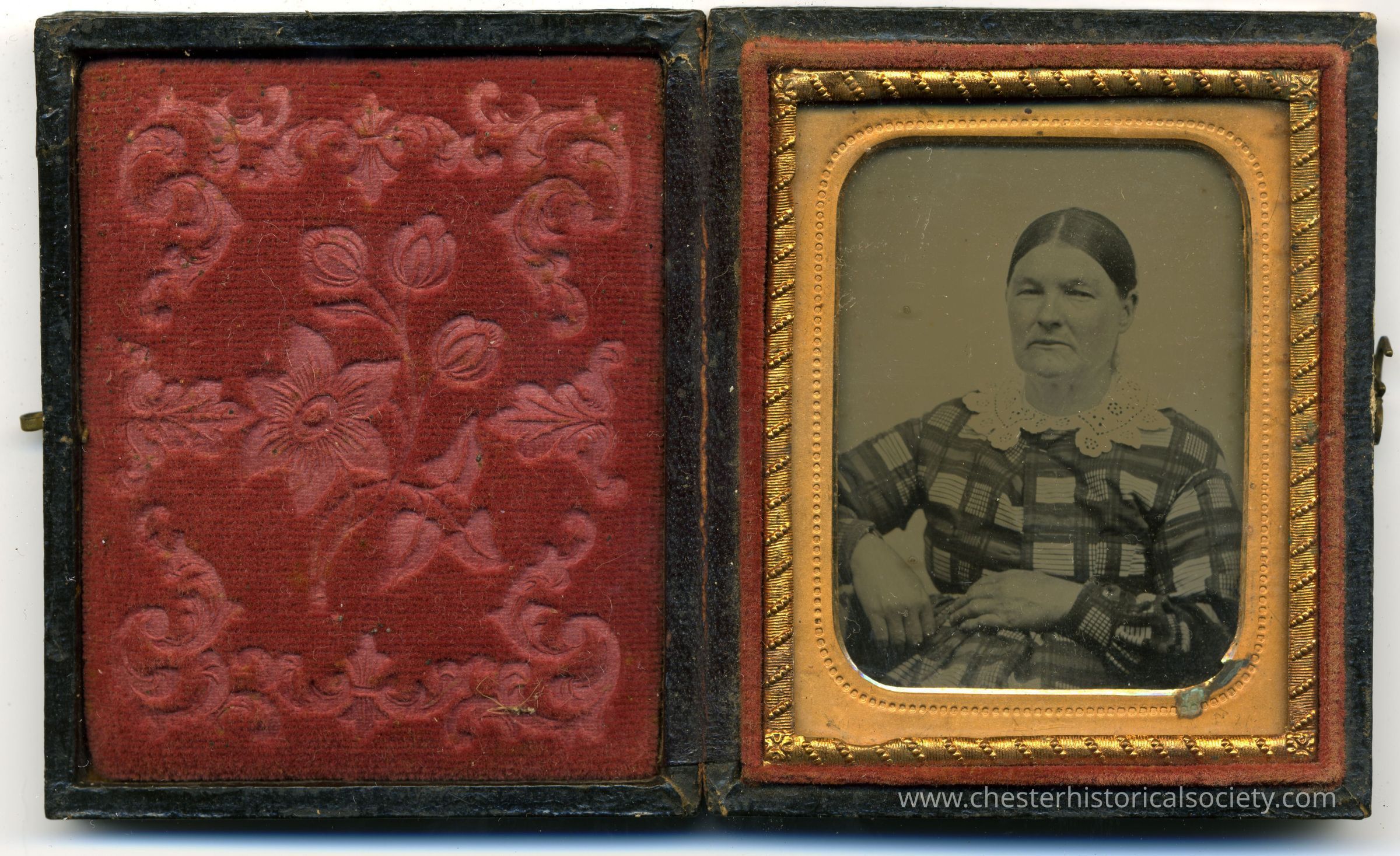 This image shows an antique photograph housed in a hinged, dual-sided case: one side displays a plush red fabric with a floral pattern, and the other side reveals a daguerreotype portrait of an elderly woman wearing a dark checkered dress adorned with a lace collar, her hands gently resting in her lap, framed by an ornate gold border.