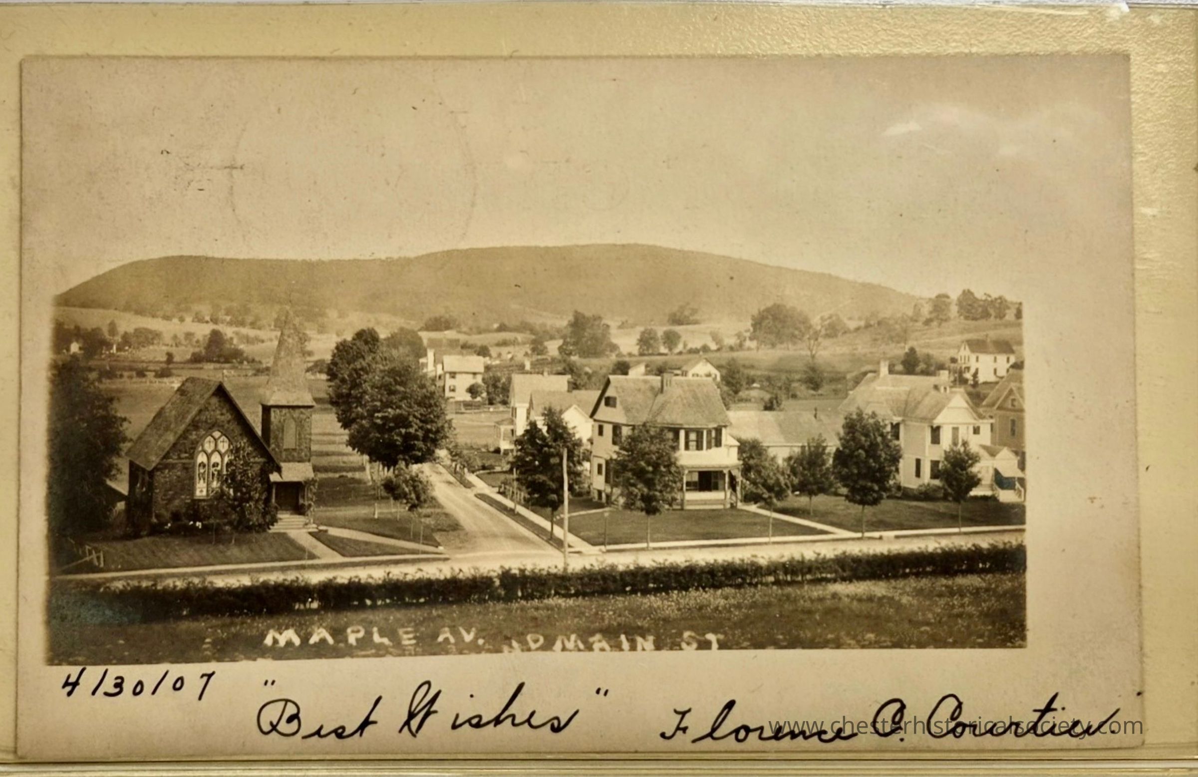 The image is an early 20th-century black and white photograph of a quaint, rural town captured on April 30, 1907. The scene features a charming view of a small community nestled against a backdrop of rolling hills under a clear sky. A prominent building in the foreground appears to be a church, distinguished by its tall, pointed steeple and ornate arched windows. Adjacent are several well-kept homes lined with lush trees, adding to the picturesque setting. A dirt road runs through the center, flanked by greenery, guiding the eye towards the distant landscape. The words “Maple Av. and Main St.” are visible, identifying the location. Handwritten at the bottom are the phrases “Best Wishes” and the signature “Florence C. Courtier,” suggesting the photo may have been sent as a postcard or keepsake. This historical snapshot offers a glimpse into the serene and idyllic life of an early American town.