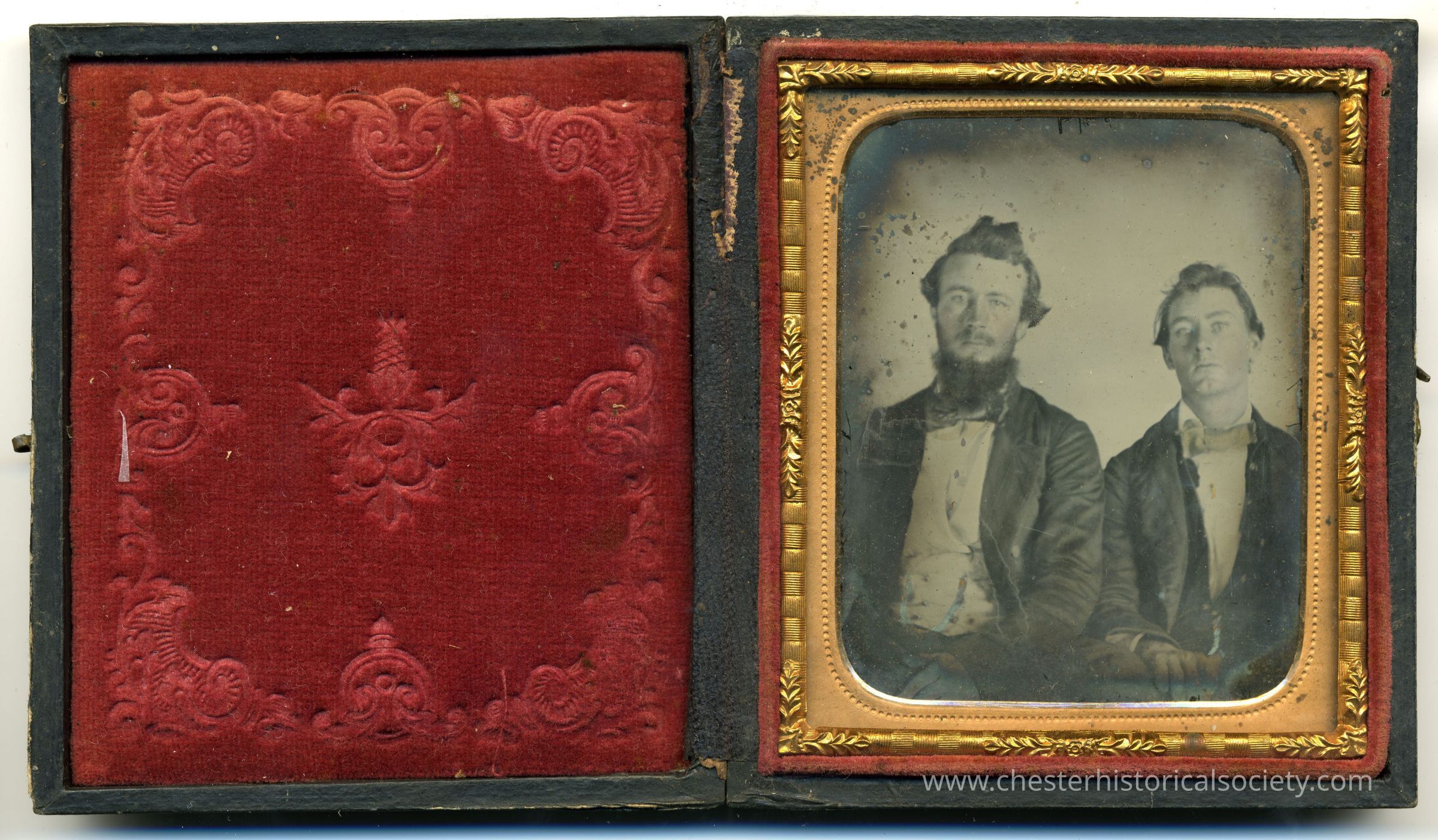 This image shows an open antique case made of black leather, which is lined with a red velvet fabric featuring ornate floral patterns. Inside the case is a daguerreotype, protected by a glass cover and framed with a gold gilded border. The photograph depicts two men seated side by side; both appear dressed in mid-19th-century attire, with the man on the left sporting a beard and the other displaying a clean-shaven look, imparting a historical glimpse into the era's fashion.