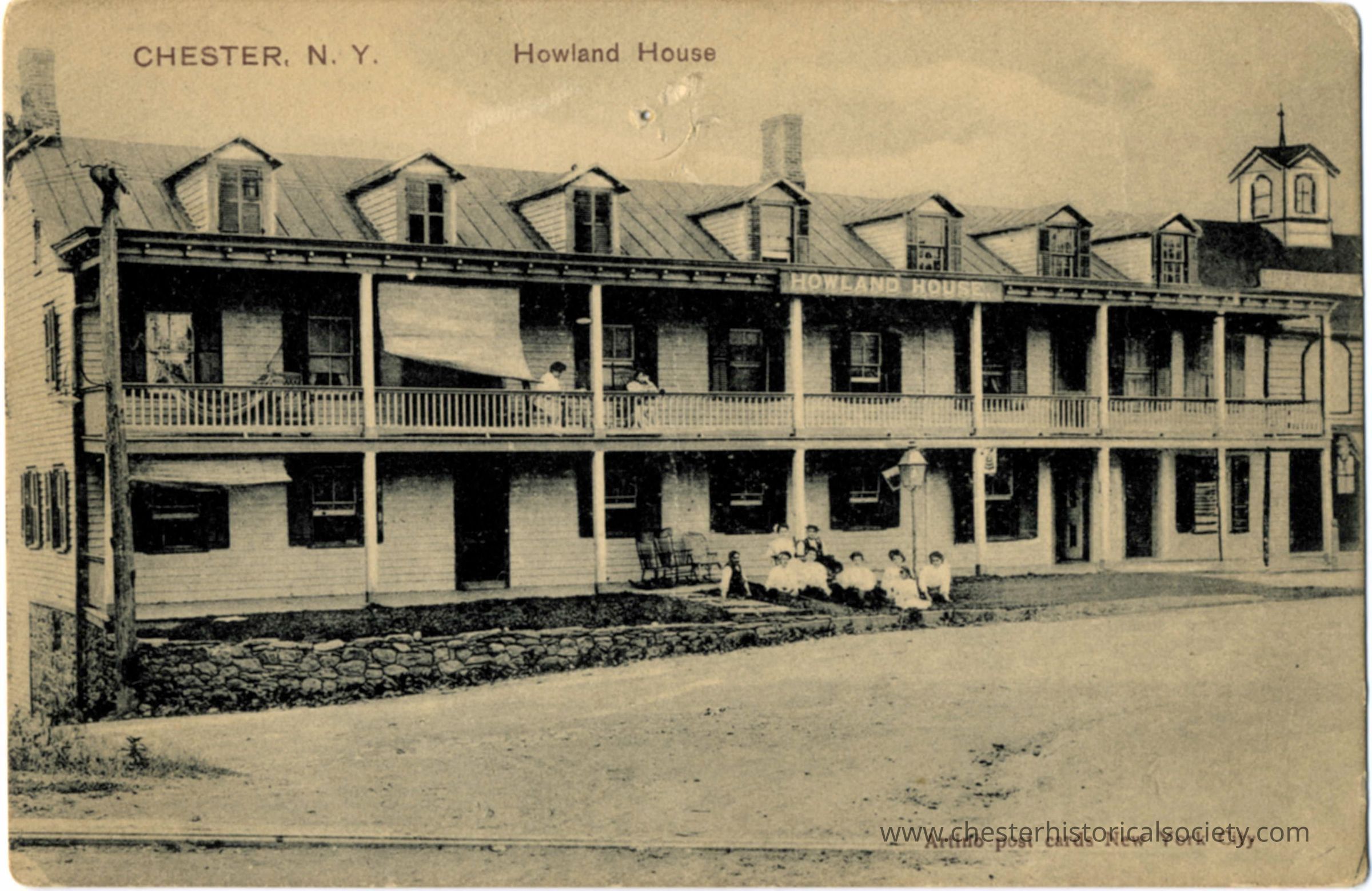 Howland House, Chester, N. Y.: This monochrome photo postcard displays the Howland House in Chester, New York, showing a large two-story wooden building with a covered porch running across both levels. There are several people, mostly women in dresses, congregating on the lawn and balconies, lending an air of social activity to the setting. The building appears aged but charismatic, with multiple architectural details like gabled windows and a central cupola over the livery stables.