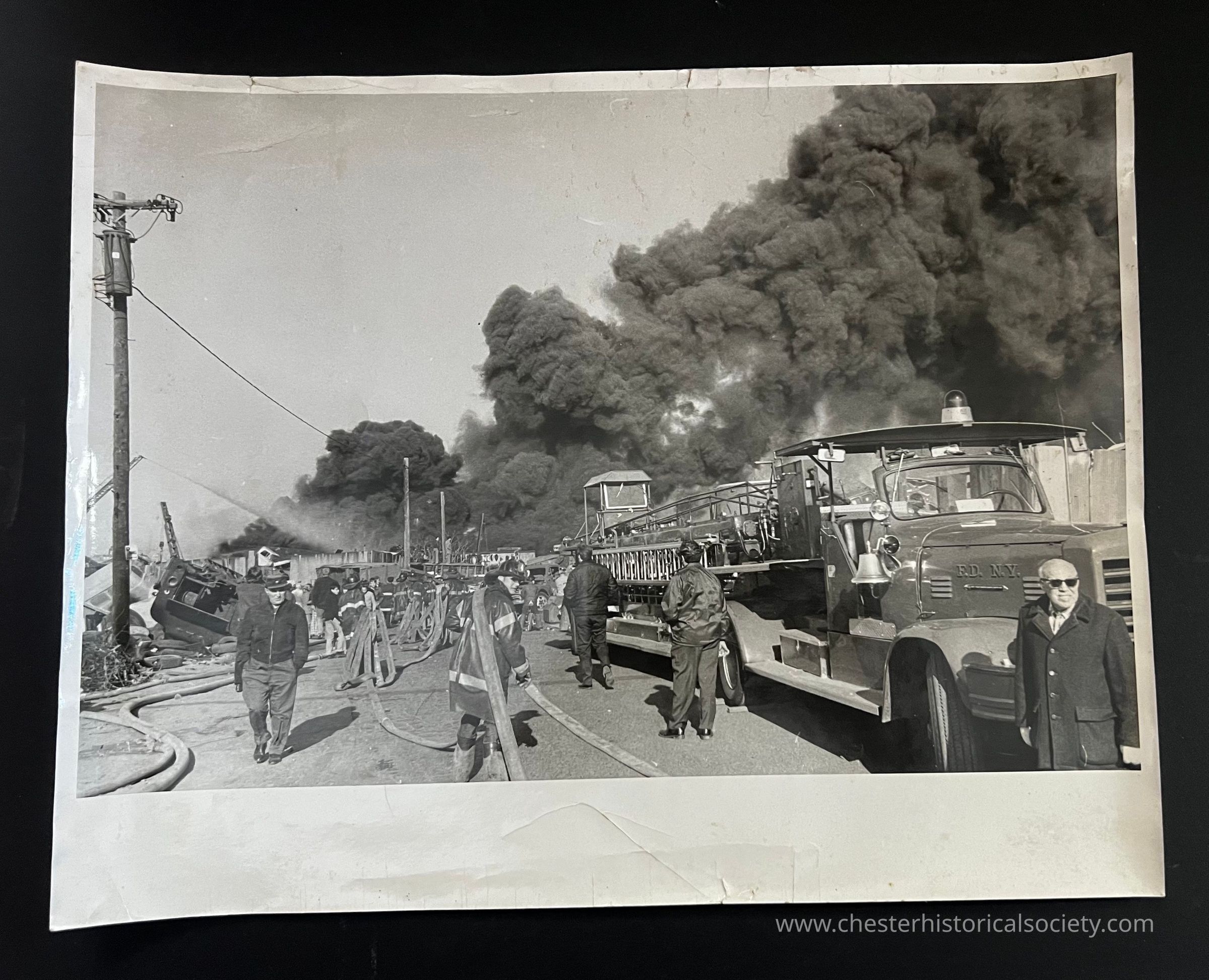 In this black and white photograph, a dramatic scene unfolds as firefighters and personnel respond to a massive fire, indicated by the dense plumes of dark smoke billowing into the sky from a distance. Fire trucks, prominently visible on the right, are stationed with hoses laid out across the ground, suggesting an active firefighting effort. Individuals, some dressed in uniforms and others in civilian attire, are seen moving purposefully, likely coordinating efforts amid the chaos. The atmosphere is tense and urgent, with the thick smoke.
