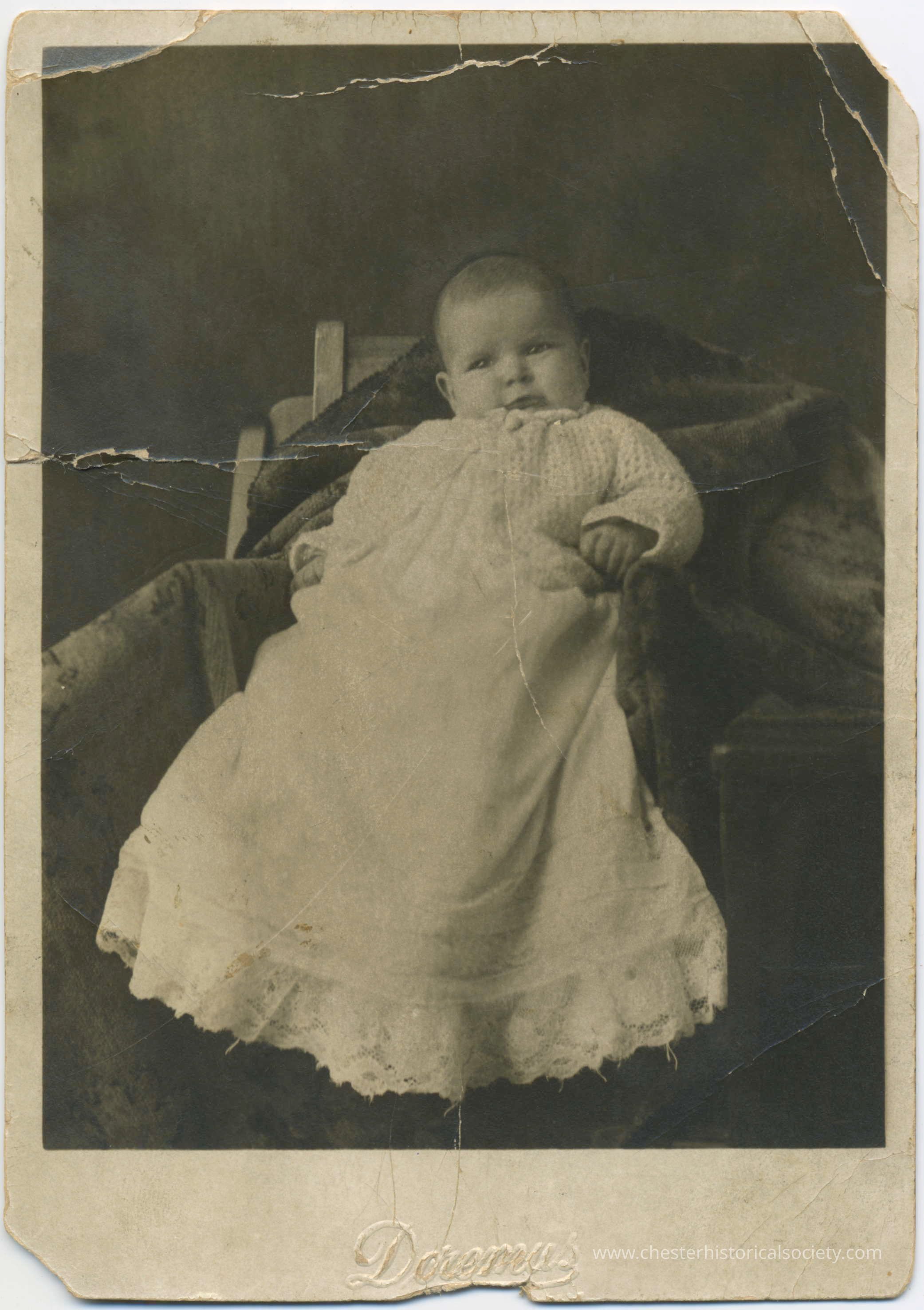 Marion McMunn baby photo postcard: This photo postcard presents a vintage, black-and-white portrait of a baby dressed in a long, lace-trimmed gown and knitted sweater, appearing serious while reclining on an ornate, fabric-draped chair. The photograph, marked with the name "Doremus" embossed at the bottom, is worn and damaged, featuring numerous cracks and tears, conveying the age and historical significance of the captured moment.