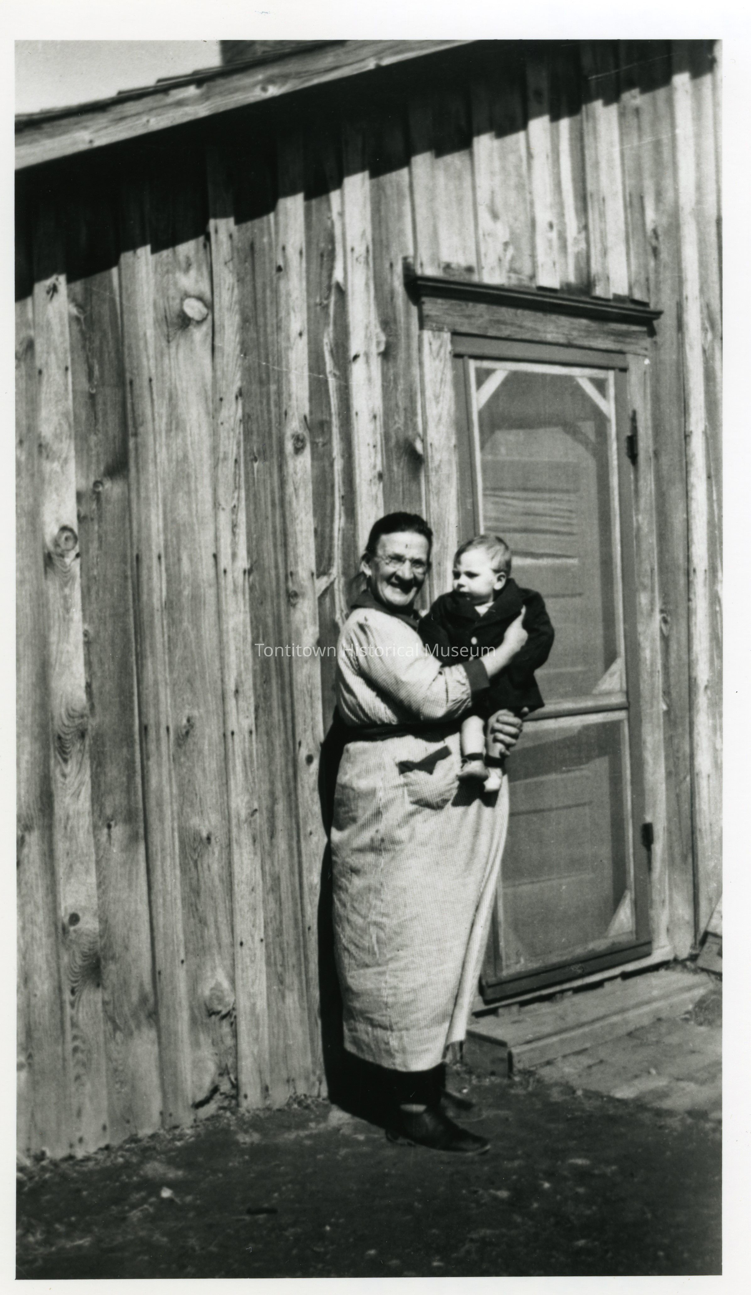 In this black and white image, an elderly woman is standing in front of a wooden house, holding a baby in her arms and smiling warmly; she is wearing an apron, and the rustic wooden exterior of the house serves as a backdrop, complementing the nostalgic and timeless feel of the photo, with shadows cast on the ground indicating a sunny day, while the baby in the woman's arms gazes curiously ahead, both figures embody a sense of familial love and connection through generations, capturing a precious moment
