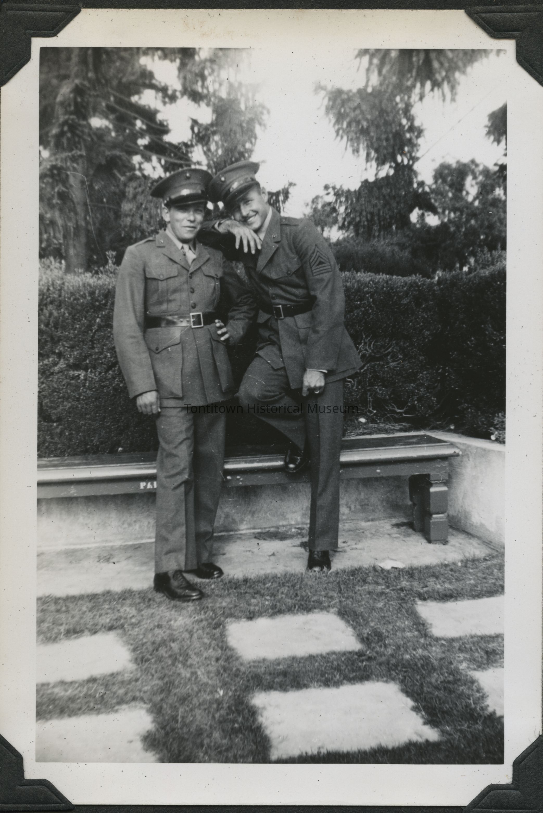 Two men in military uniforms stand together for a photo.: In the black-and-white photograph, two men in military uniforms and caps stand closely together, exuding camaraderie and a sense of unity; the man on the right casually rests his arm on the shoulder of the man on the left, who stands with one hand on his hip, both smiling slightly and appearing relaxed against the backdrop of a garden with neatly trimmed hedges and tall trees in the distance.