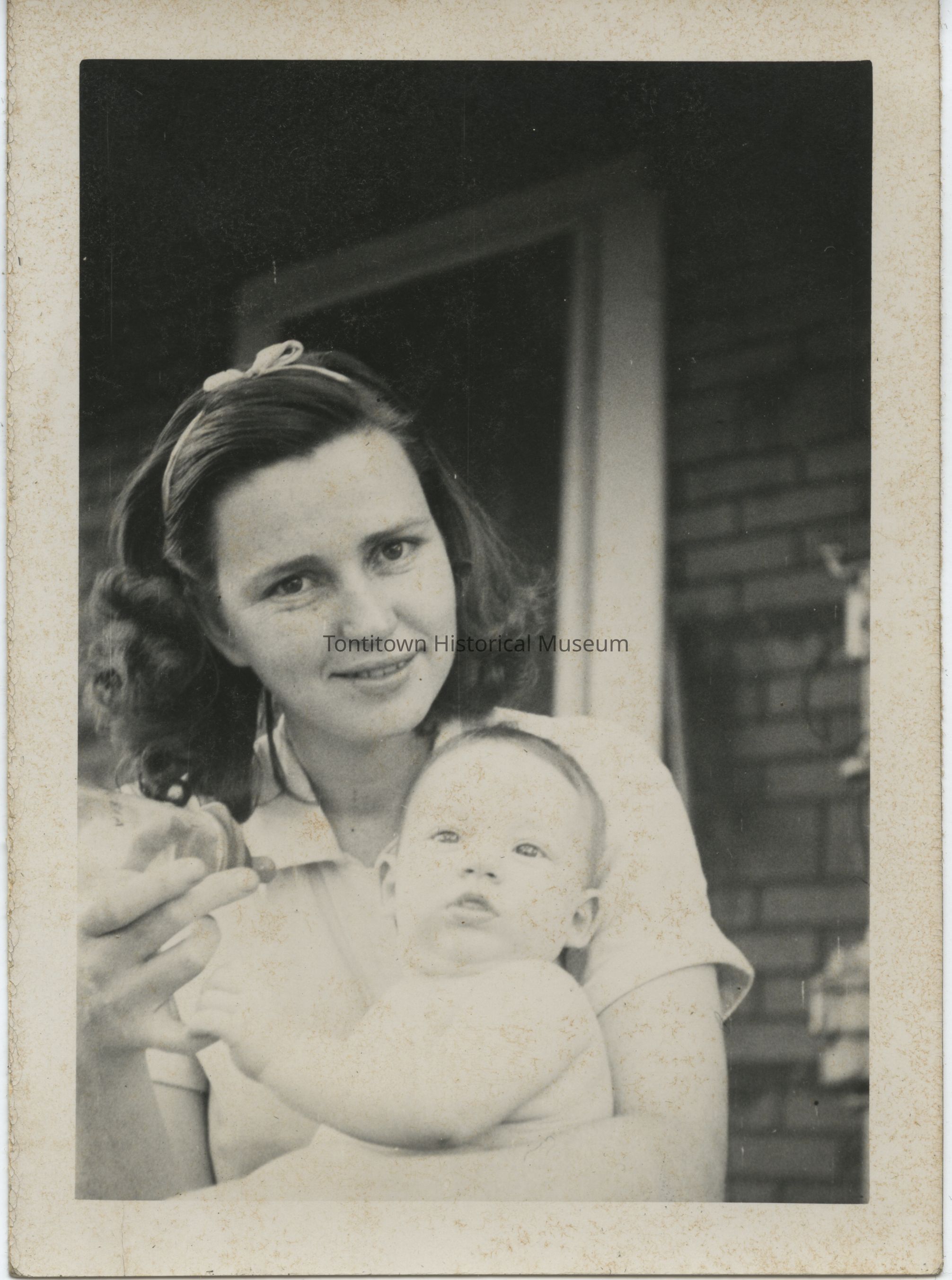 Woman with baby smiling at the camera.: In the sepia-toned photograph, a woman with curled hair and a headband lovingly cradles an infant in her arms; the baby, gazing upwards with an inquisitive expression, is held securely while she tenderly offers a milk bottle, suggesting a moment of affectionate care and nurturing, set against a backdrop of a brick building.