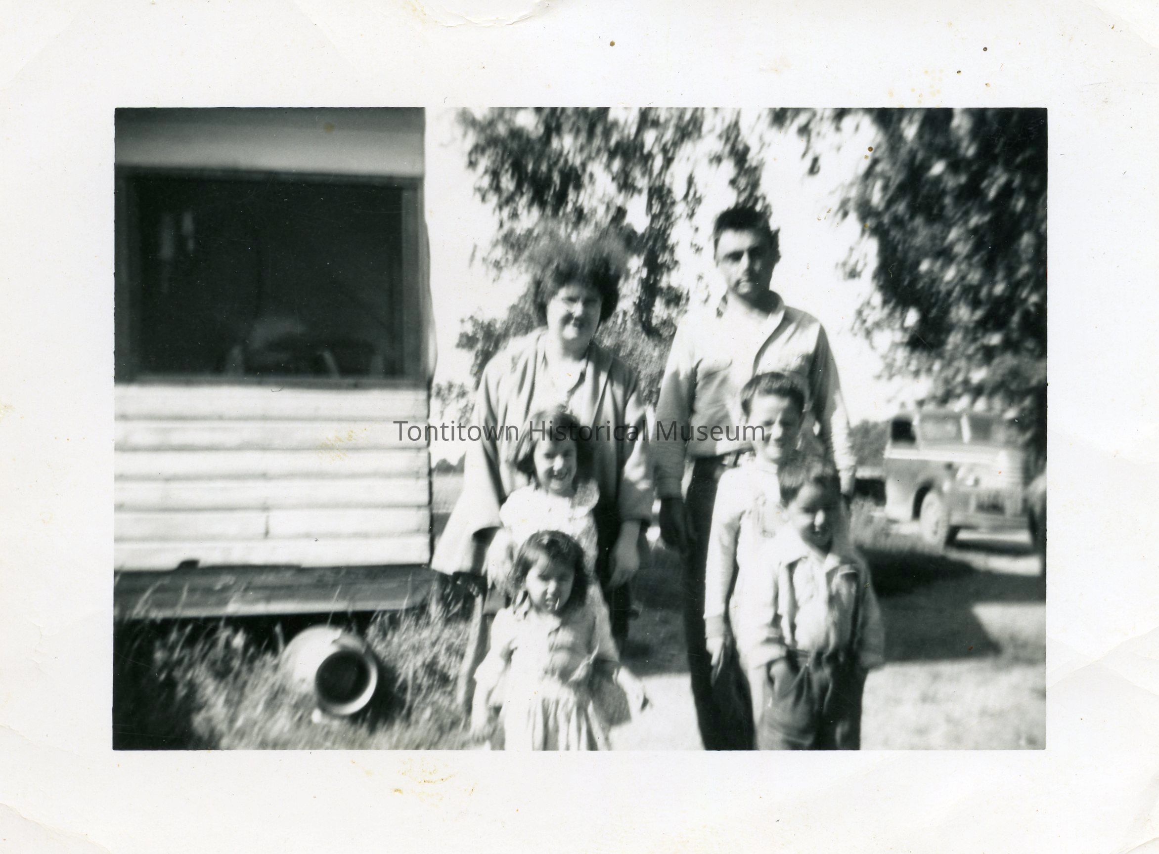 A vintage black-and-white photograph captures a family of six standing outdoors. In the background, there is a building partially visible, and a vehicle parked nearby under the shade of a tree. The woman and man, likely the parents, stand together behind four children who are clustered in front, dressed in typical mid-20th century attire. The setting suggests a rural or suburban area, surrounded by greenery, evoking a sense of nostalgia and capturing a moment in the family's life. The natural light enhances the