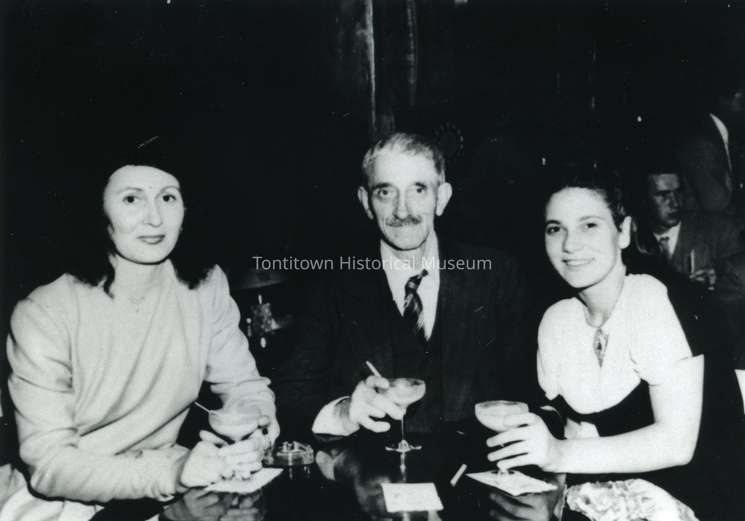 Night out on the town: The black and white image captures three individuals sitting closely together at a table in a bar or a restaurant, as indicated by the presence of cocktail glasses and an ashtray on the table; the woman on the left is wearing a light-colored dress with dark wavy hair; the man in the middle, with a mustache and wearing a suit and tie, holds a drink in hand, exuding a relaxed demeanor.