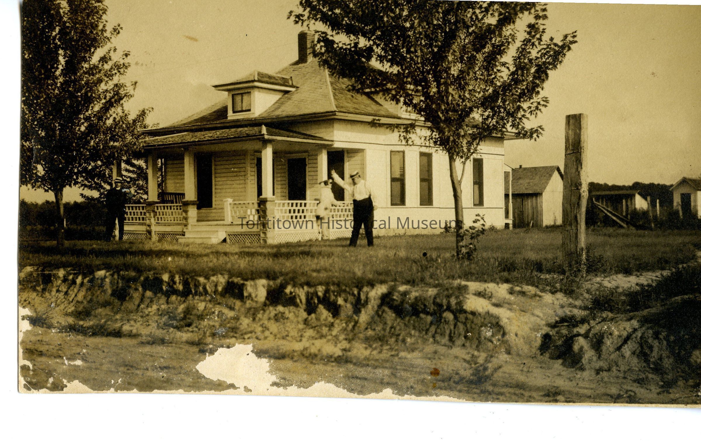 Memo Morsani, wearing dark trousers and a light shirt and a hat plays with the family dog in front of his home in Tontitown. A gentleman in a black suit and hat stands at the opposite corner of the house, which has white siding and single story with an ample front porch and attic window; out and other buildings seen in the background. View possibly from the road.